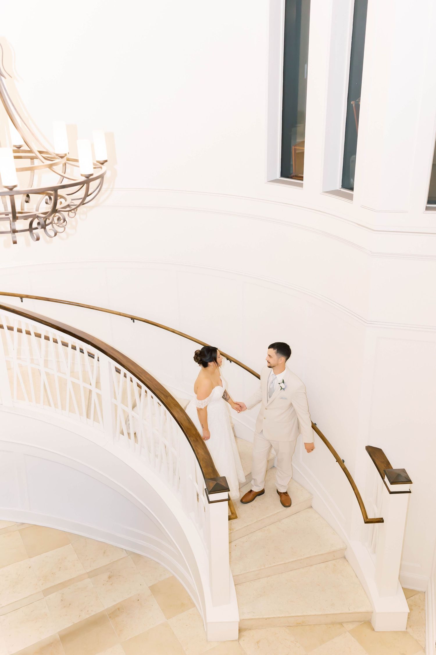 Bride and groom holding hands on elegant curved white staircase with gold railing during wedding day portraits.