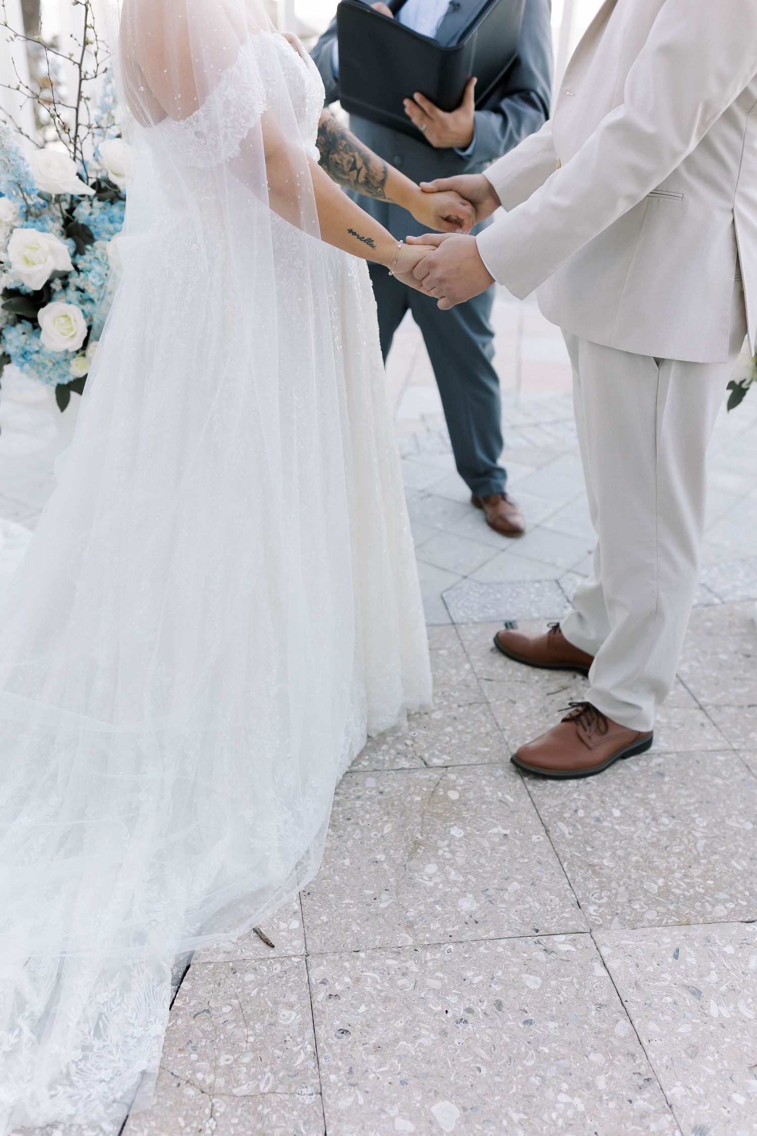 Bride and groom hold hands during outdoor wedding ceremony while officiant reads from book.