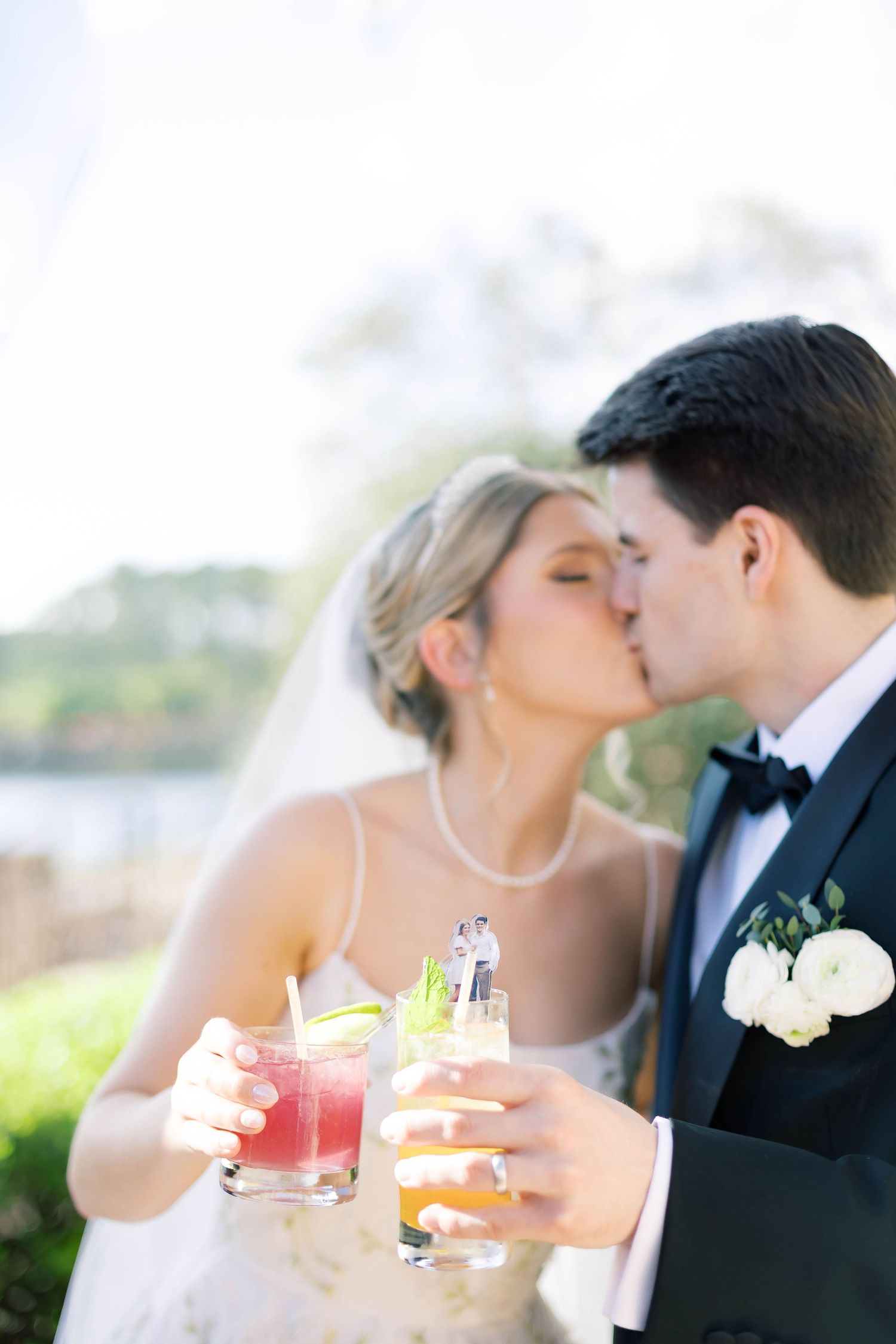 Bride and groom sharing a kiss while holding colorful cocktails outdoors on their wedding day.