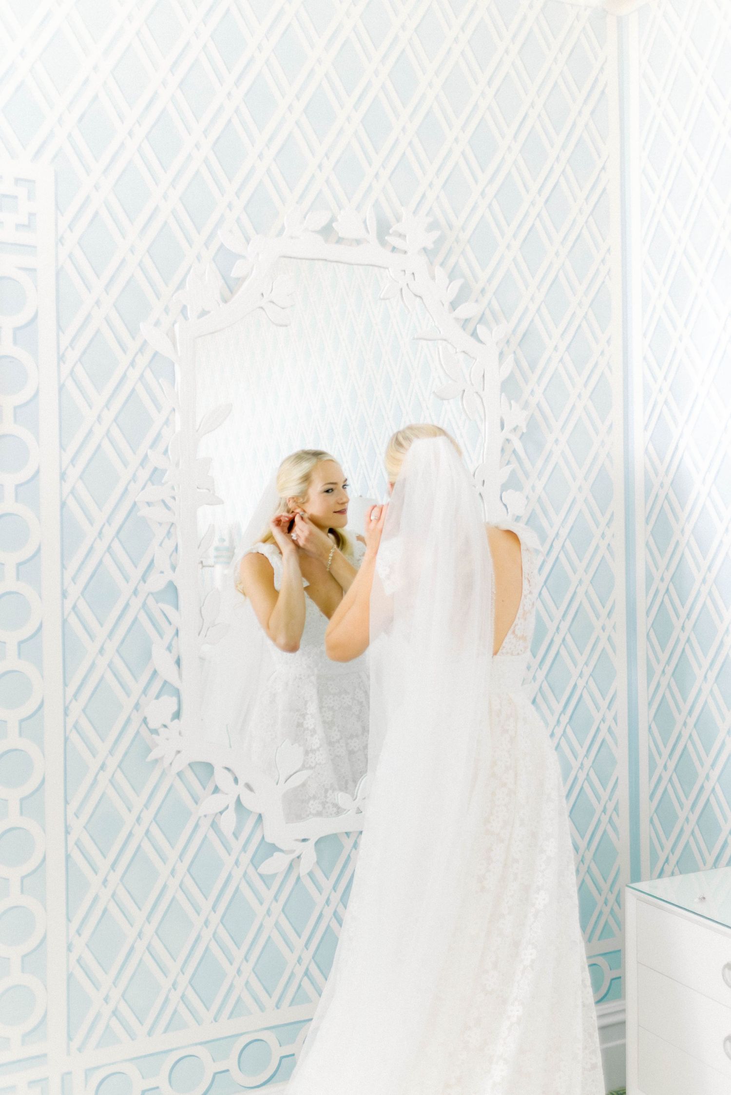 Bride in white lace gown looking at reflection in ornate mirror against blue patterned wallpaper.
