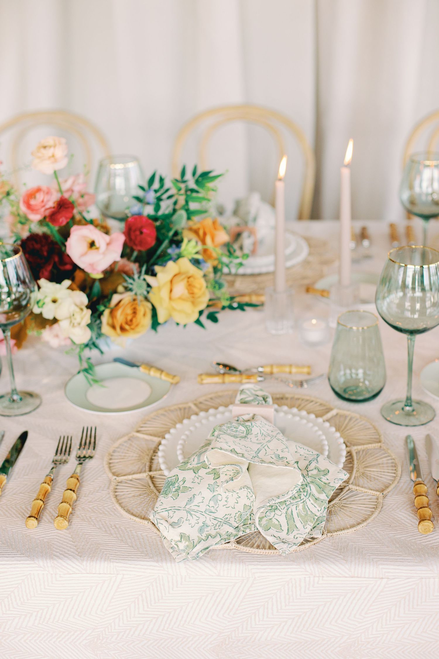 Elegant wedding table setting with gold-rimmed plates, green glassware, and colorful floral centerpieces on white linens.