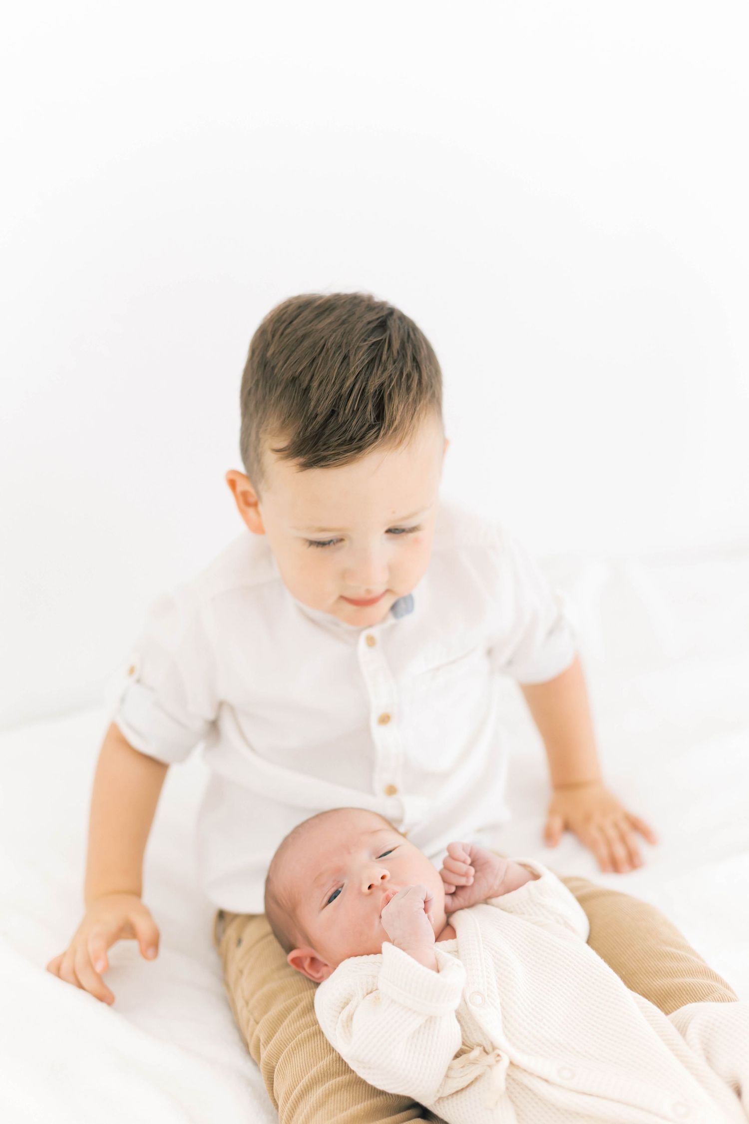Tender sibling moment with toddler in white shirt looking at newborn on white background.