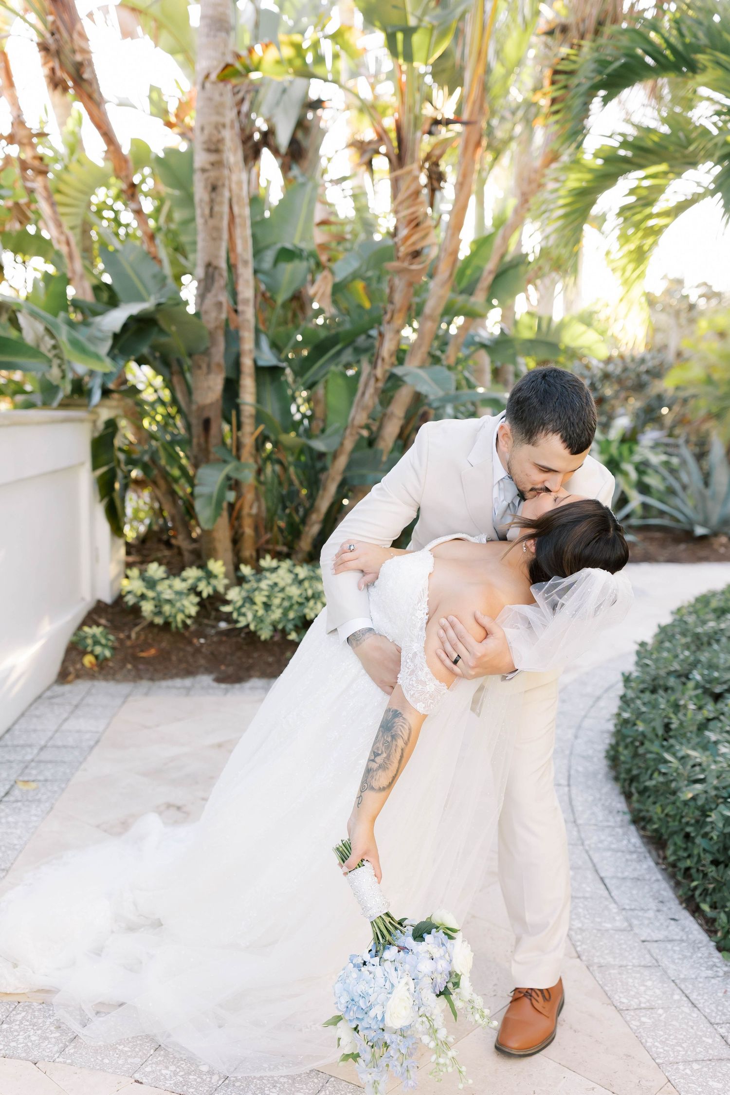 Groom dips bride for a kiss on stone pathway surrounded by tropical palms and lush greenery, bride holding blue bouquet.