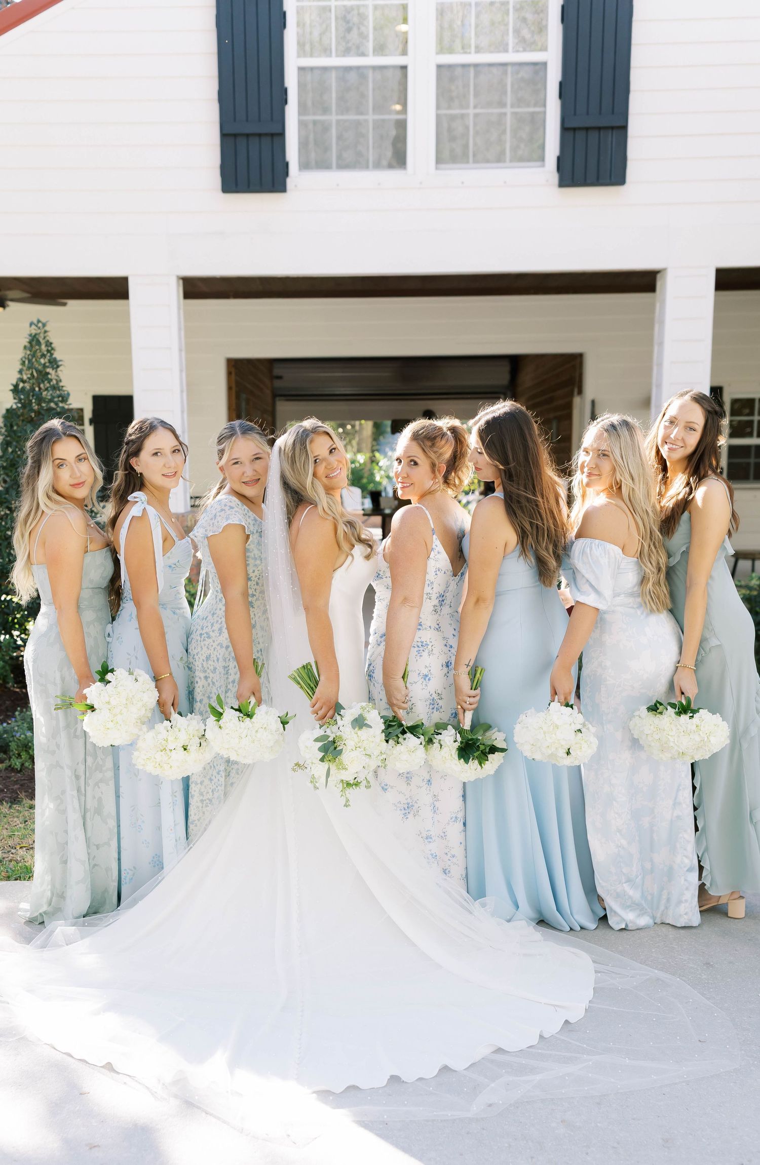 Wedding party in light blue dresses lines up against a white building with navy shutters.
