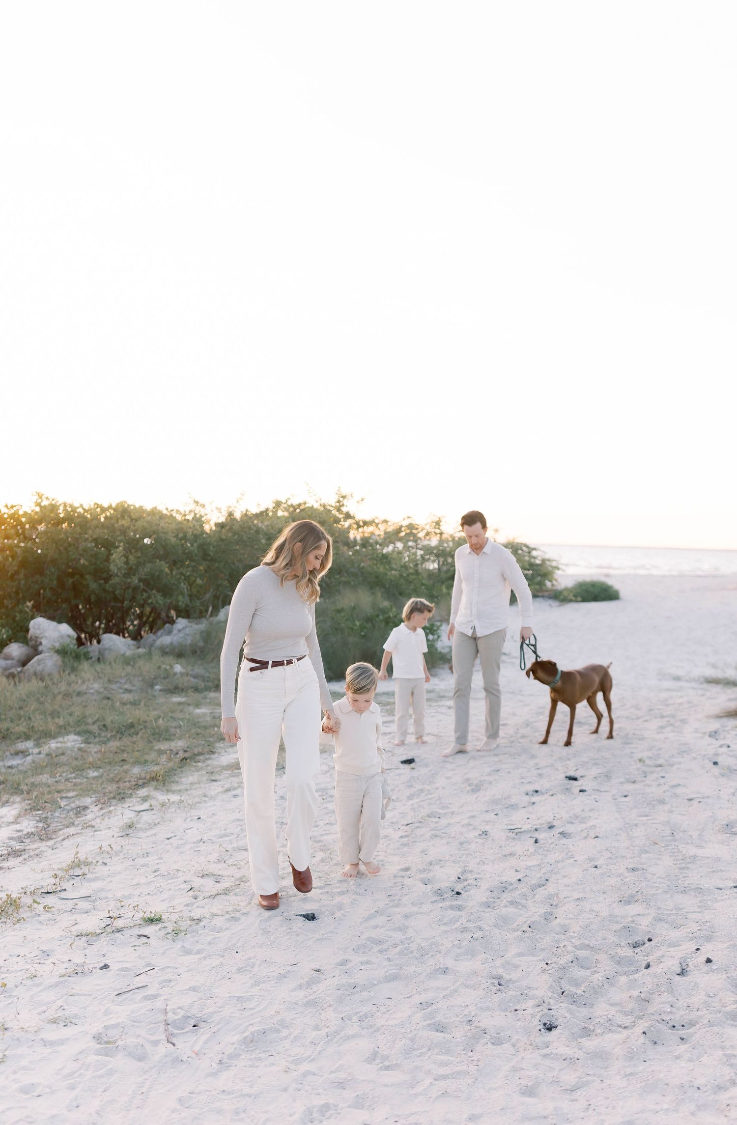 Family and their dog walk along sandy beach path in matching white attire.