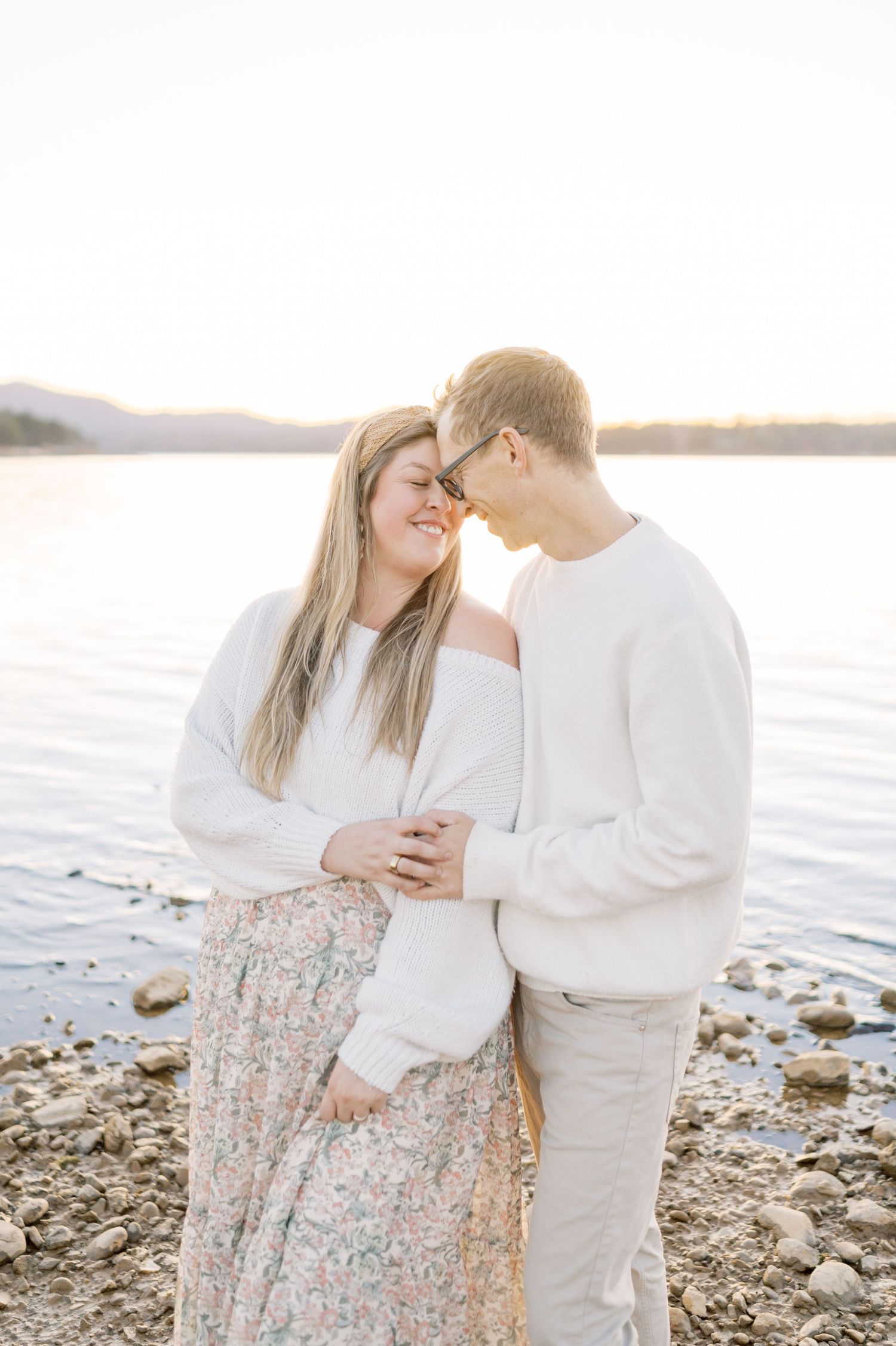 Couple embracing and smiling by a rocky lakeside at golden hour, wearing coordinating white and neutral tones.