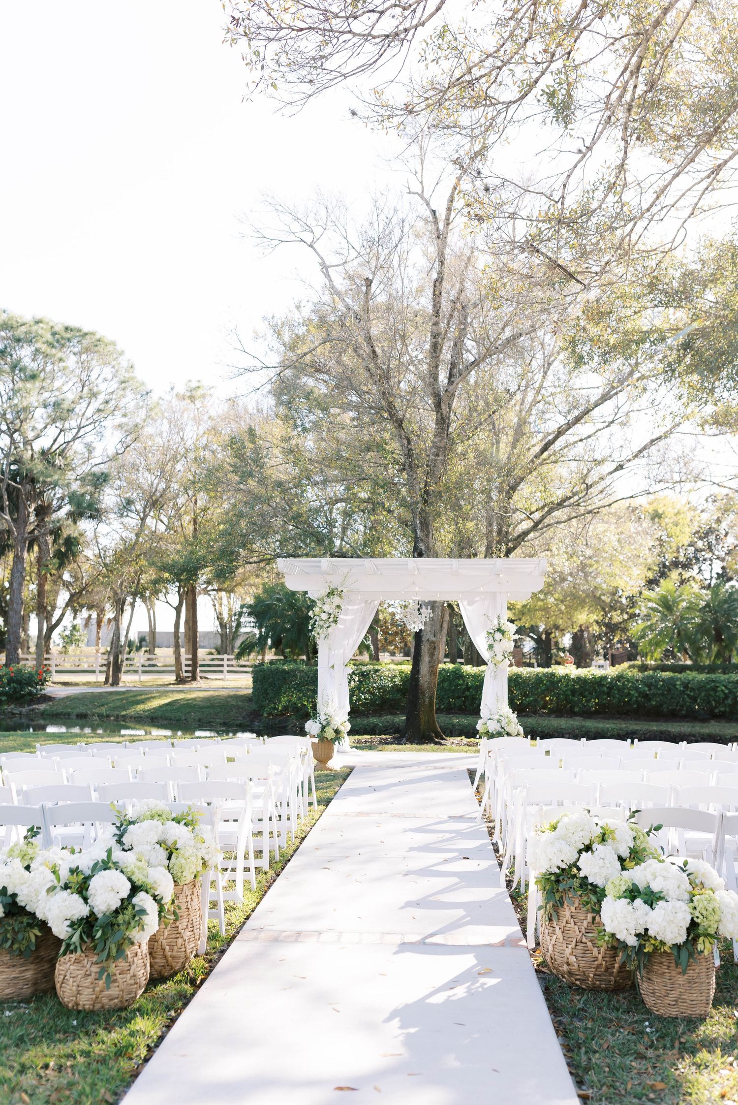 White wedding ceremony setup with woven baskets of flowers lining an outdoor aisle under trees.