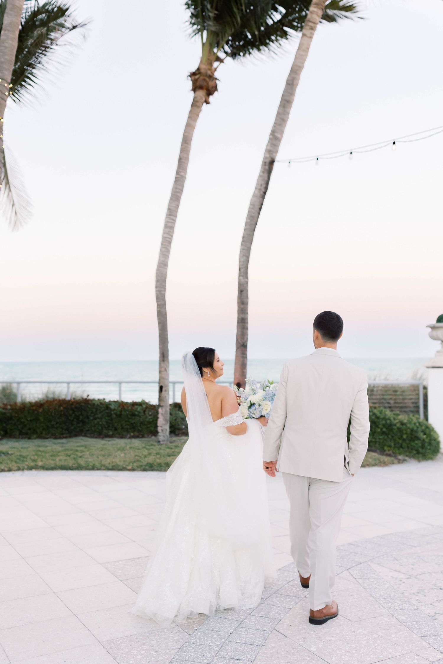 Bride and groom walk hand-in-hand toward ocean at sunset beneath tall palm trees.