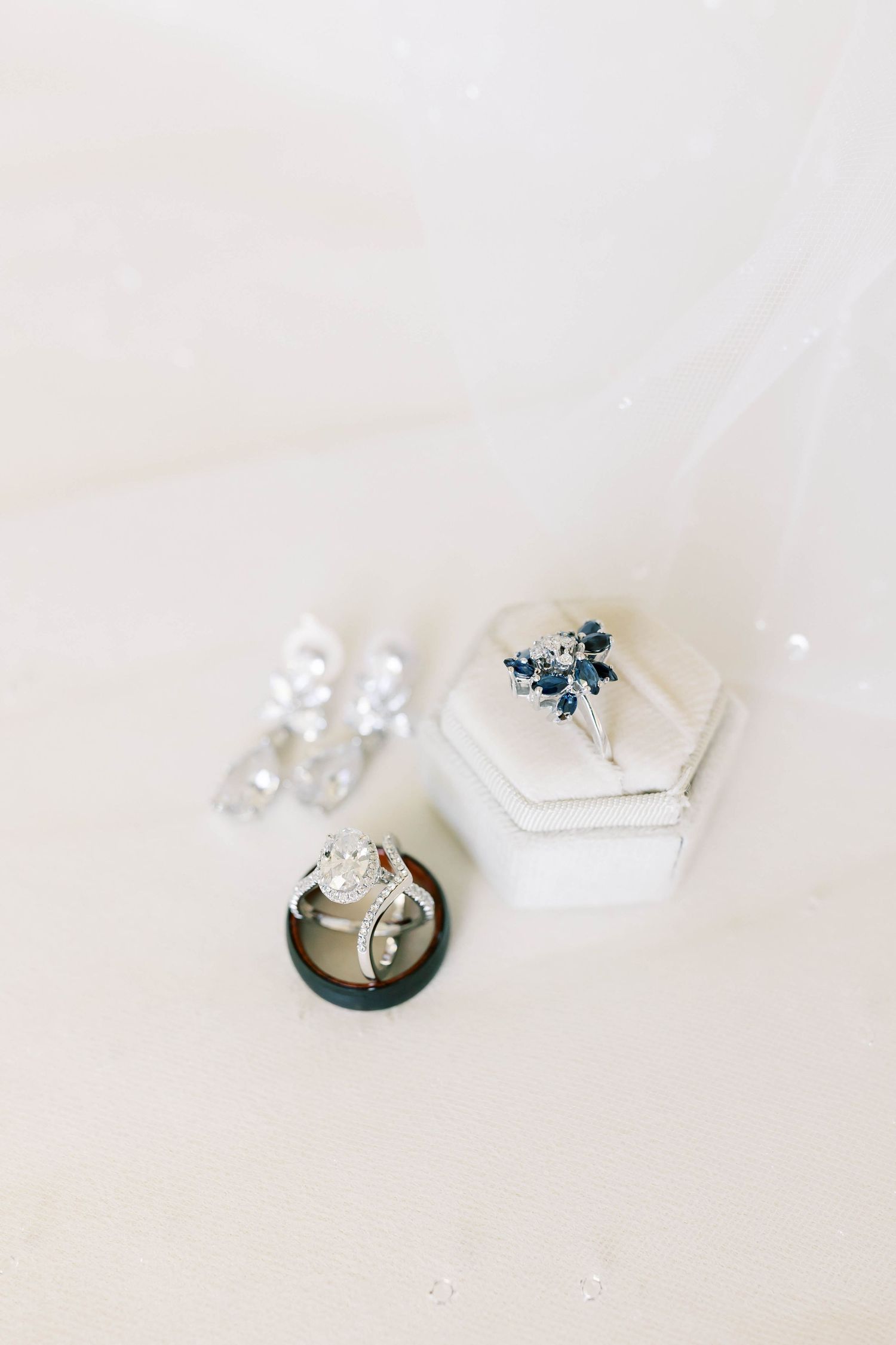 Silver diamond engagement ring and sapphire cluster ring displayed in white velvet box with crystal drop earrings on soft white background.