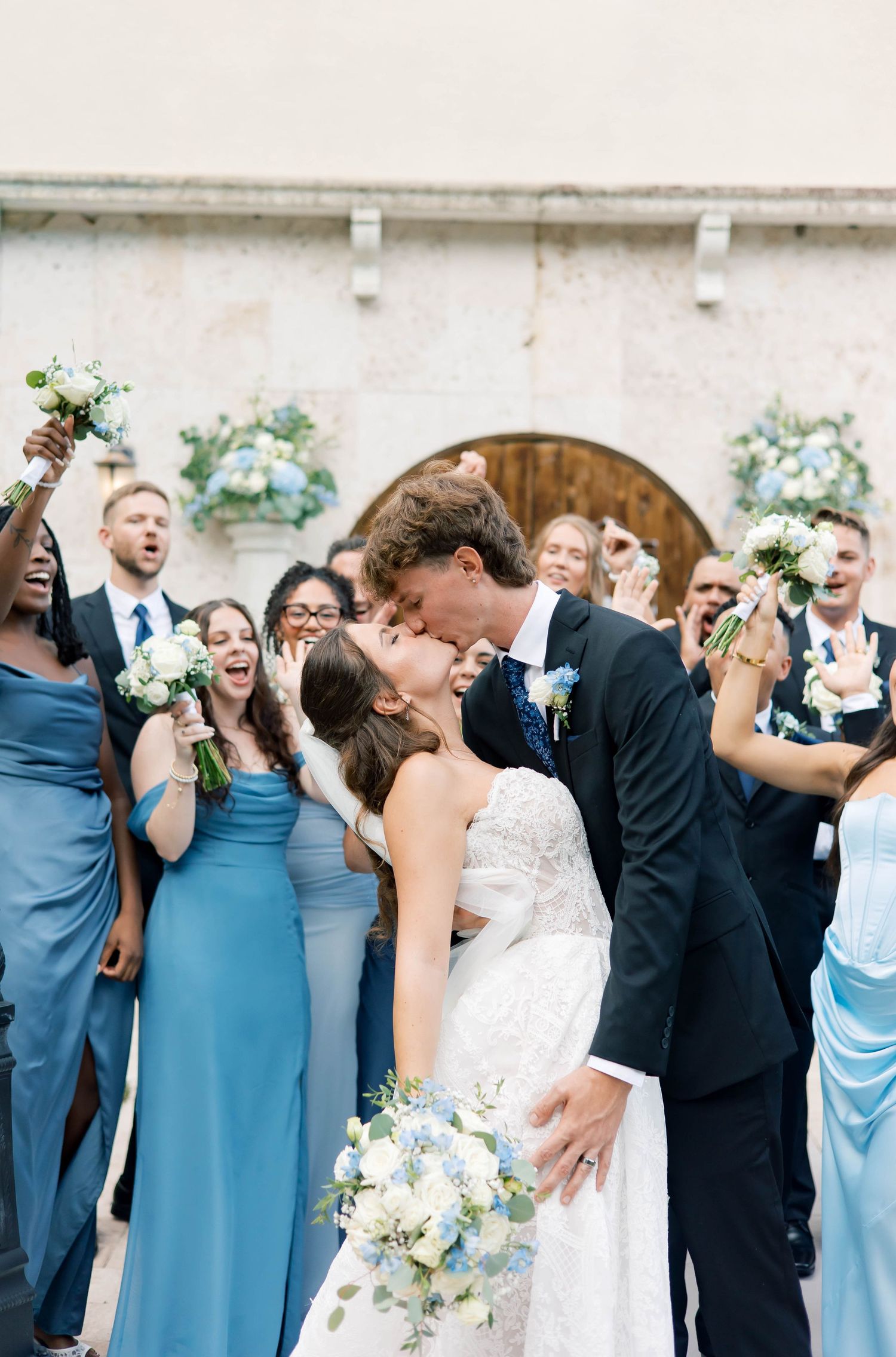 Bride and groom share a kiss surrounded by cheering wedding party in blue dresses and suits outside a church.