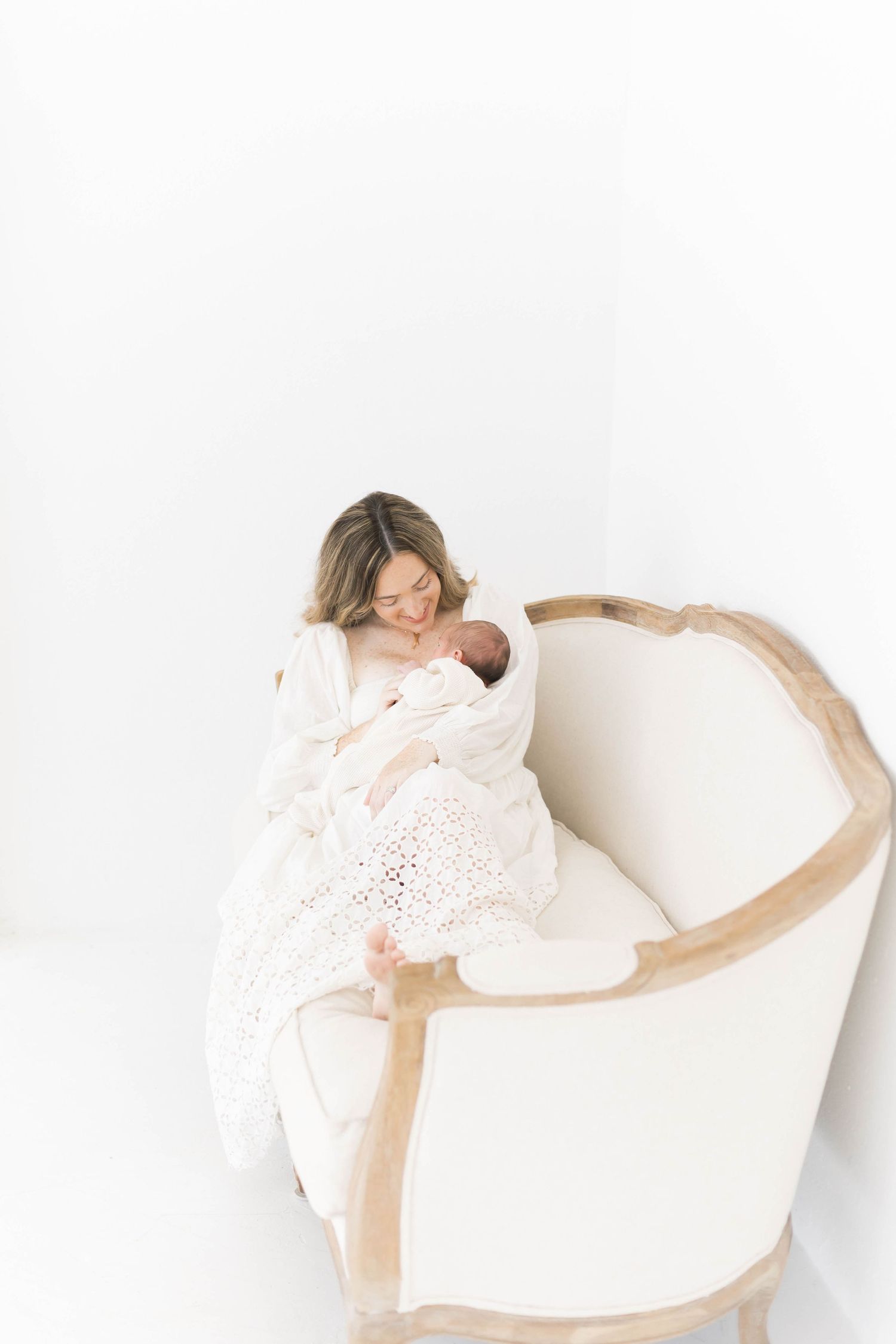 Artistic portrait of mother and newborn resting in white upholstered chair against light background.