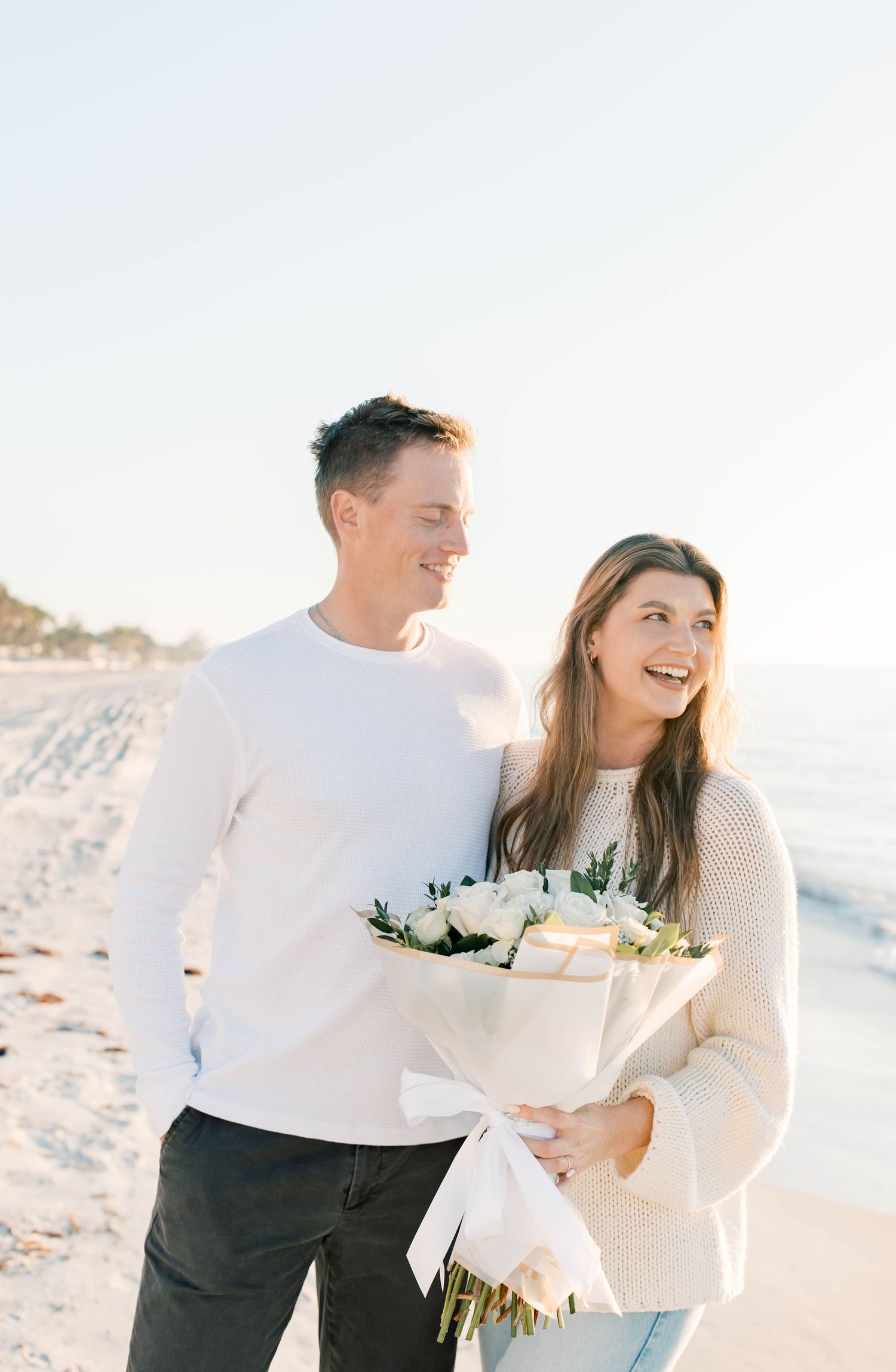 A joyful beach photoshoot featuring a couple in white holding a floral bouquet.