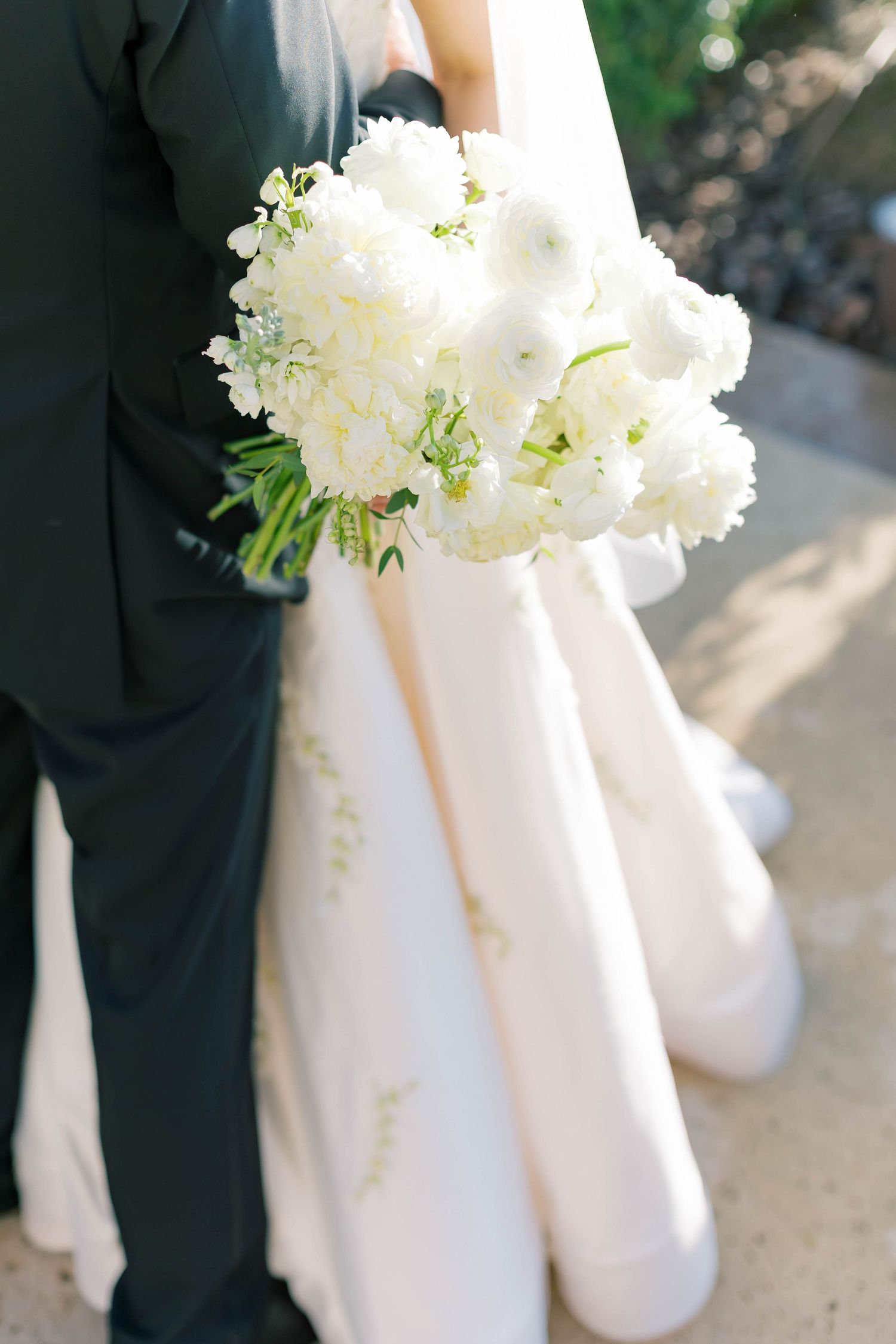 Bride in white gown holding lush white peony and ranunculus bouquet beside groom in dark suit outdoors.