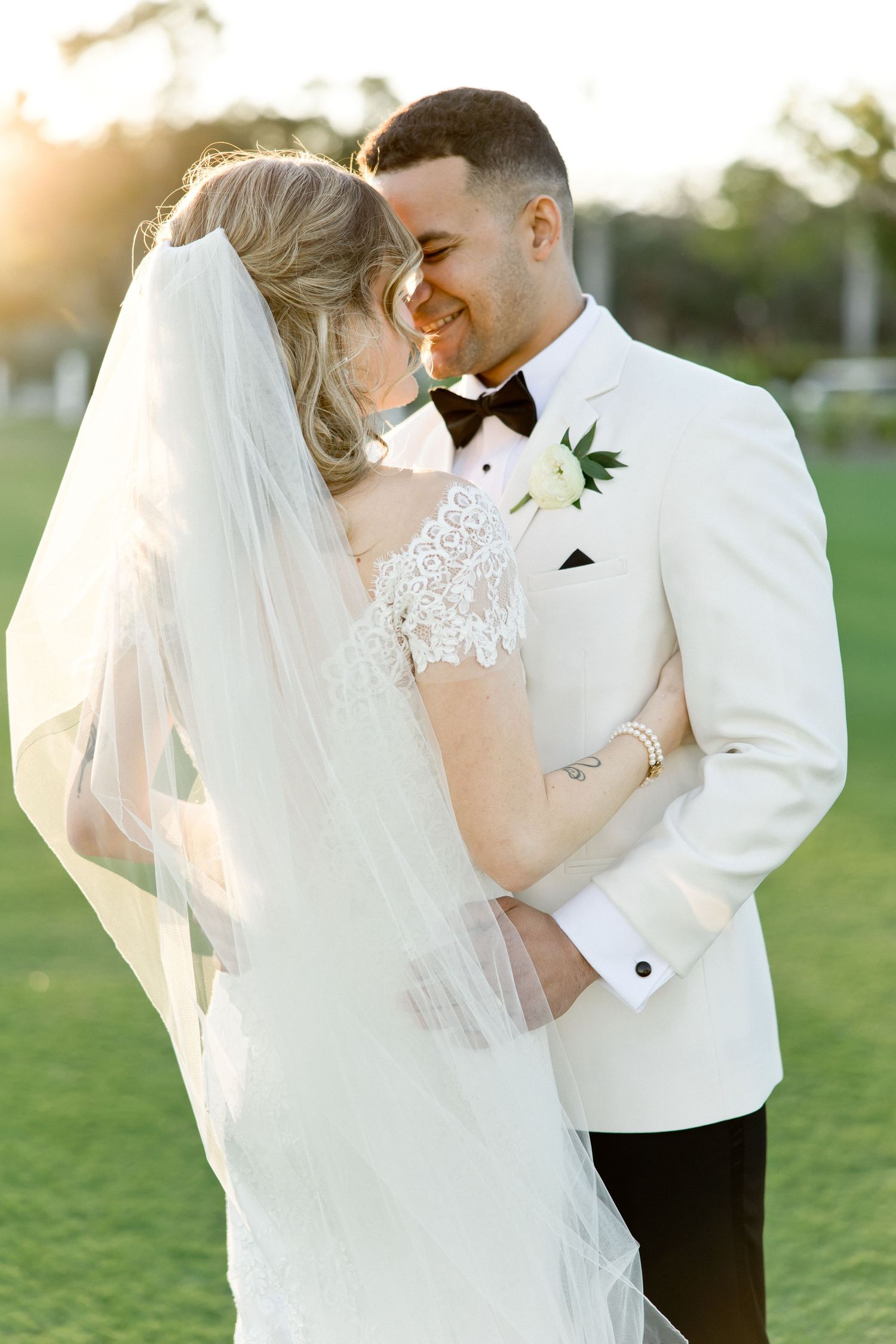 Bride and groom share a romantic moment at golden hour, with her veil flowing as they smile and embrace on a green lawn.