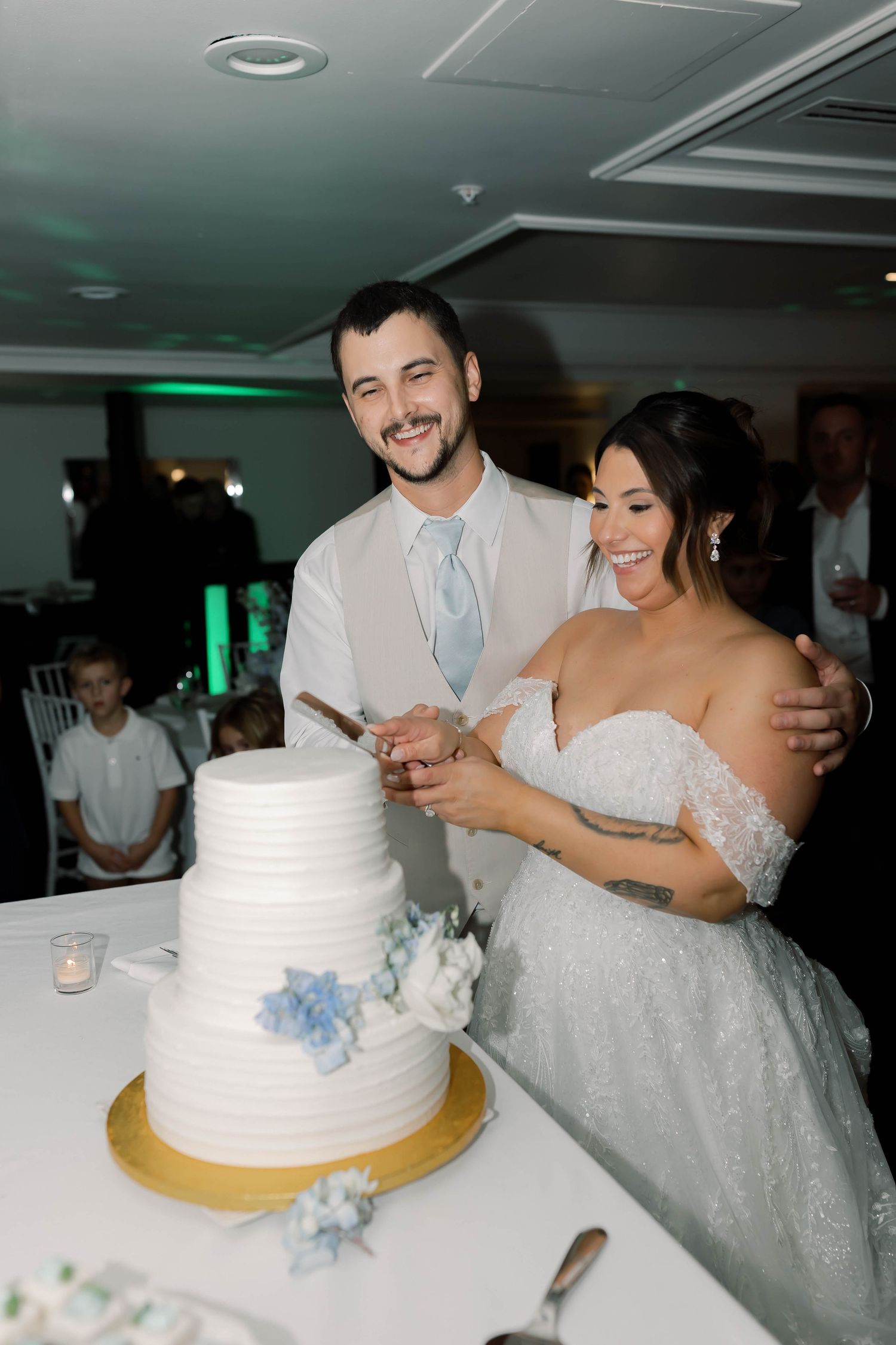 Smiling bride and groom cutting their white three-tier wedding cake with blue floral accents at their reception.