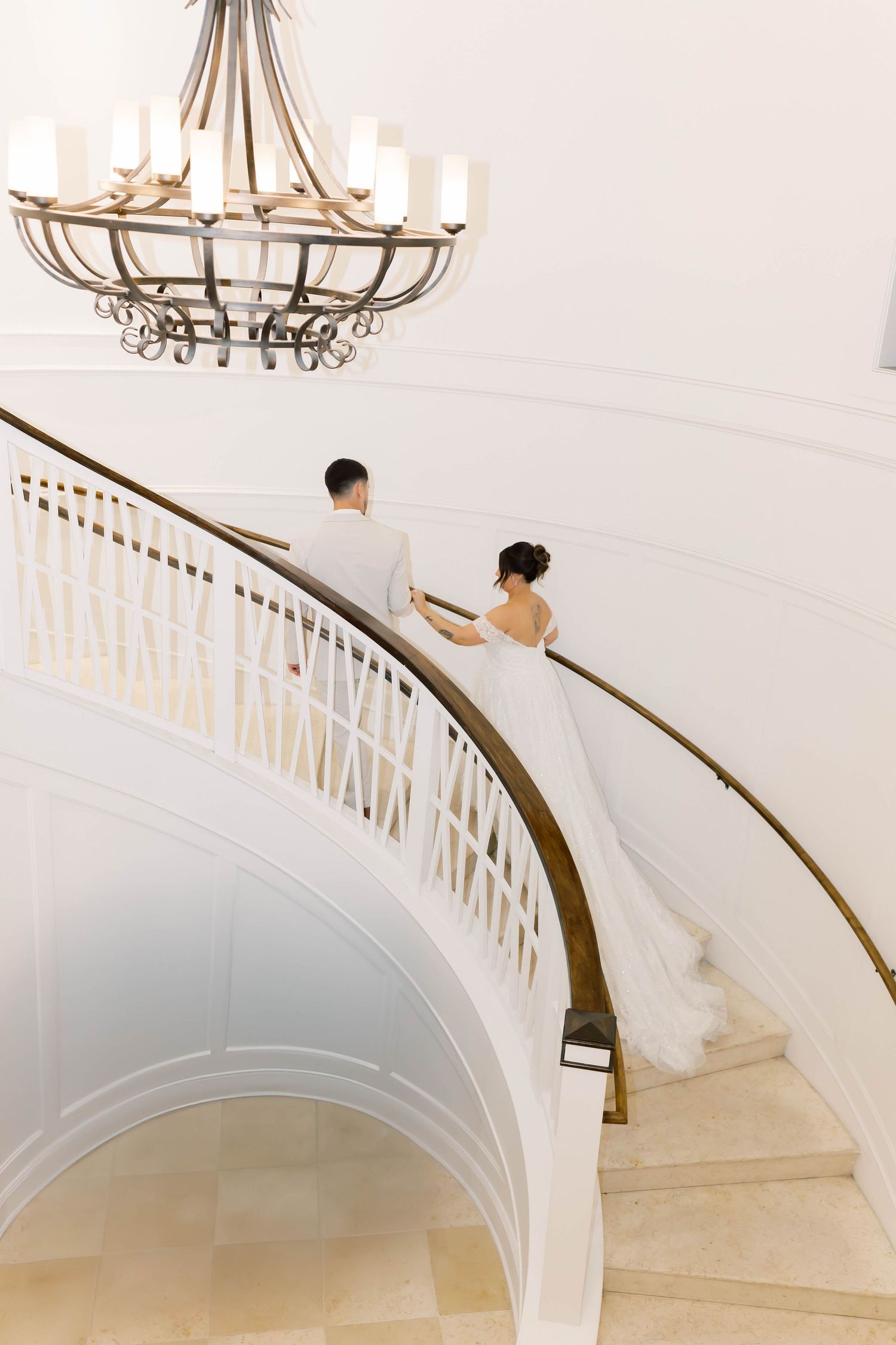 Bride and groom ascend elegant curved staircase beneath ornate chandelier in grand white interior.