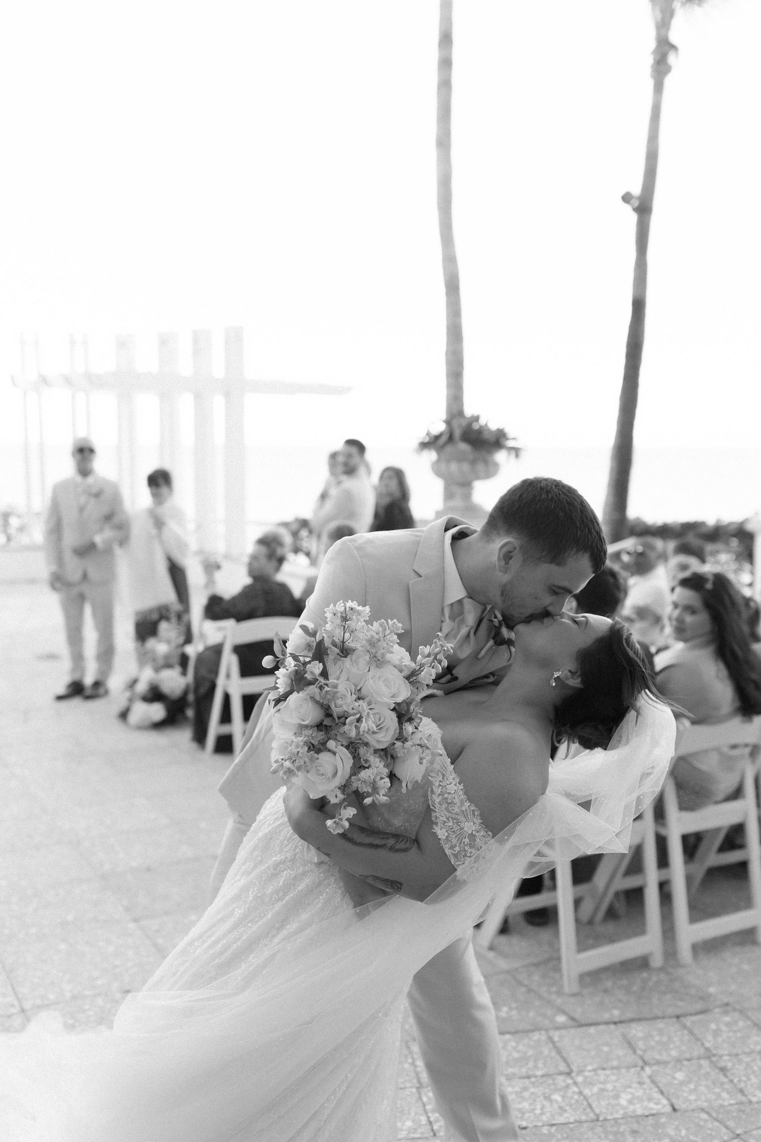 Bride and groom share a romantic dip kiss during outdoor beach wedding ceremony with guests seated in white chairs.