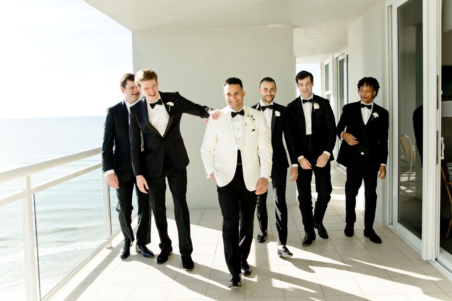 Groomsmen in formal attire standing in line on balcony overlooking ocean.