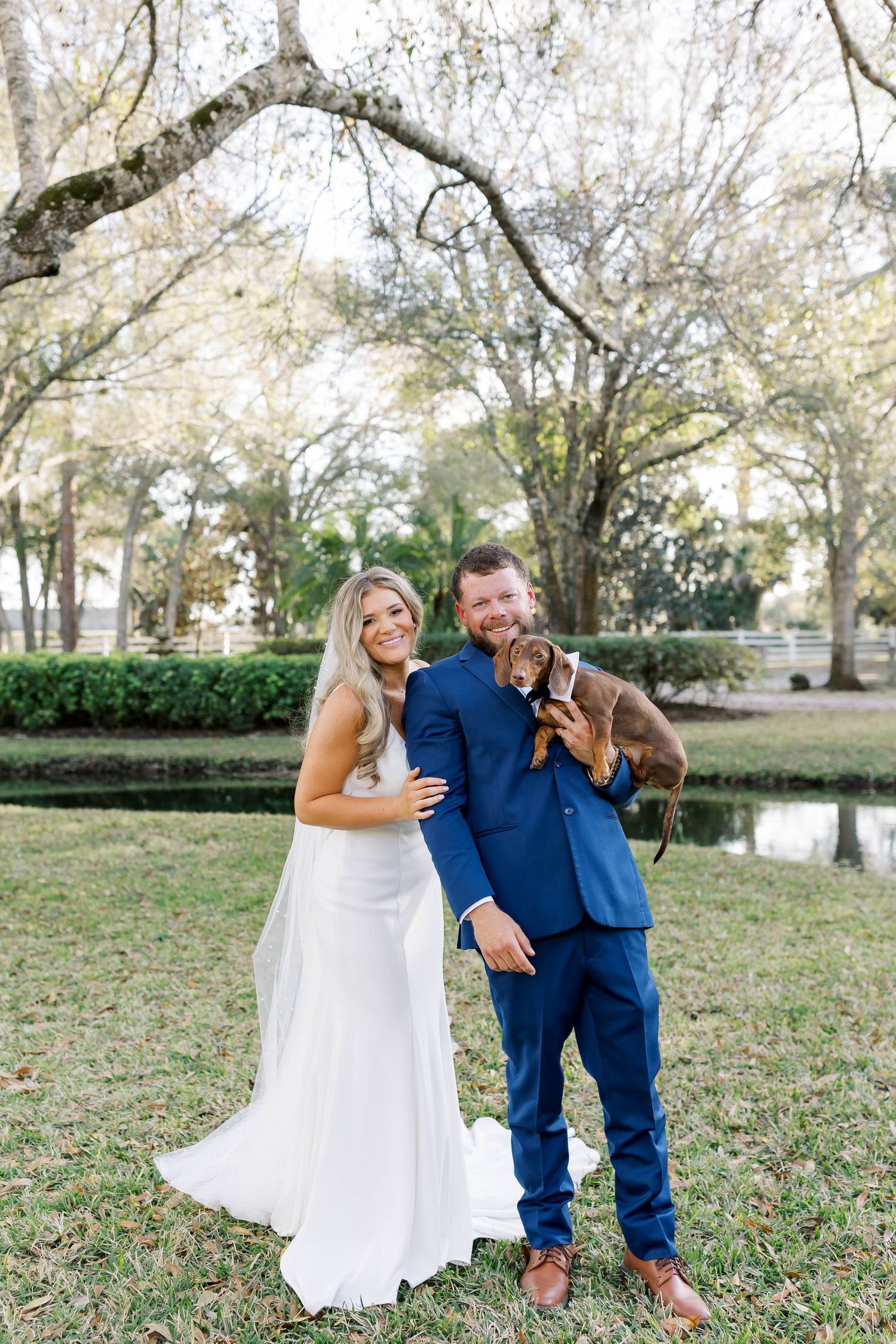 Wedding couple poses with dachshund on grassy lawn near lake.