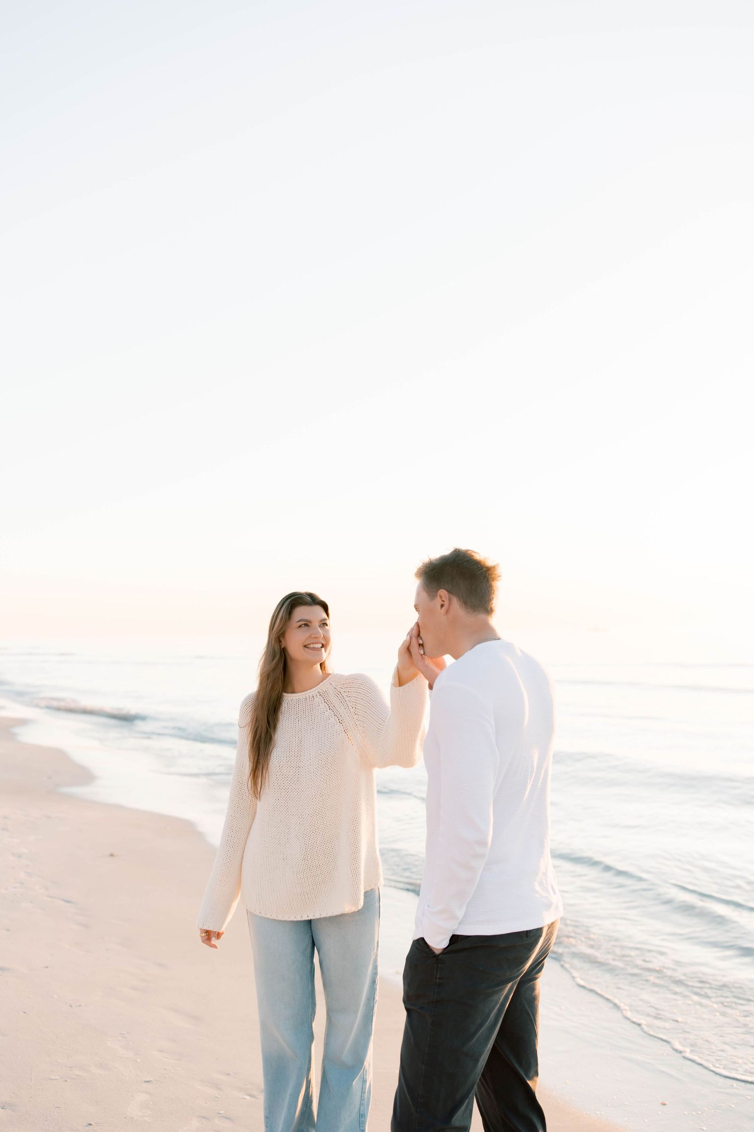 Couple in white and dark clothing walking together on a sunny beach at sunset.