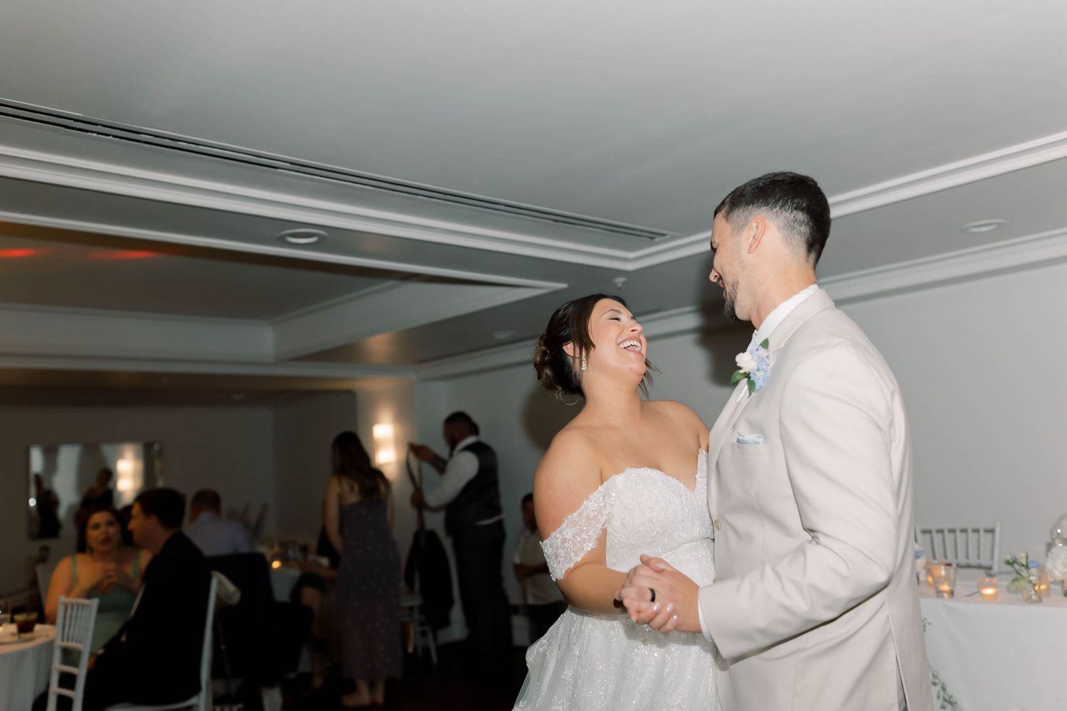 Bride in off-shoulder gown and groom in tan suit share their first dance at wedding reception.