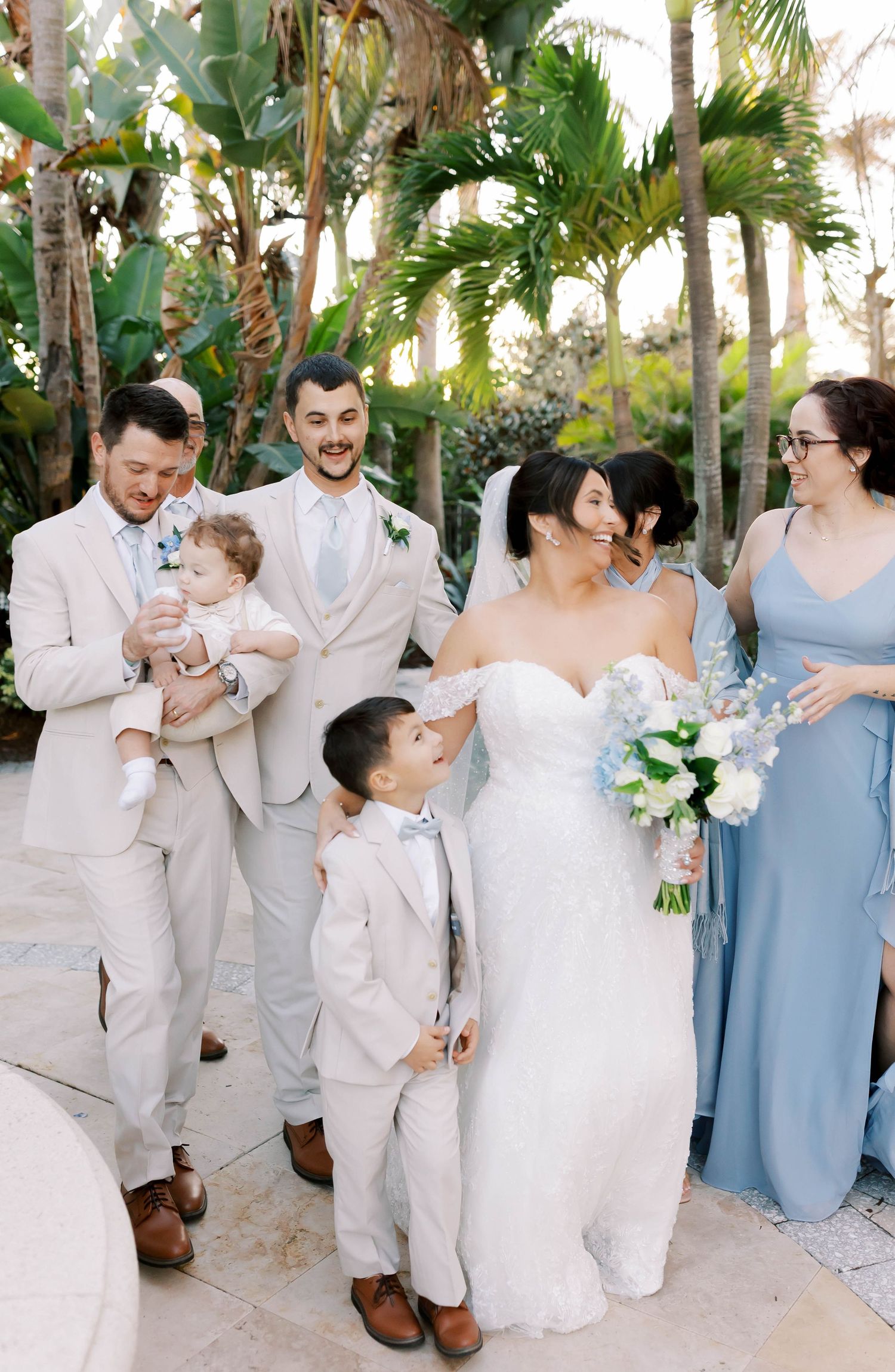 Wedding party walks outdoors in tropical setting, bride in white gown with bouquet, groomsmen in tan suits, bridesmaid in blue dress.
