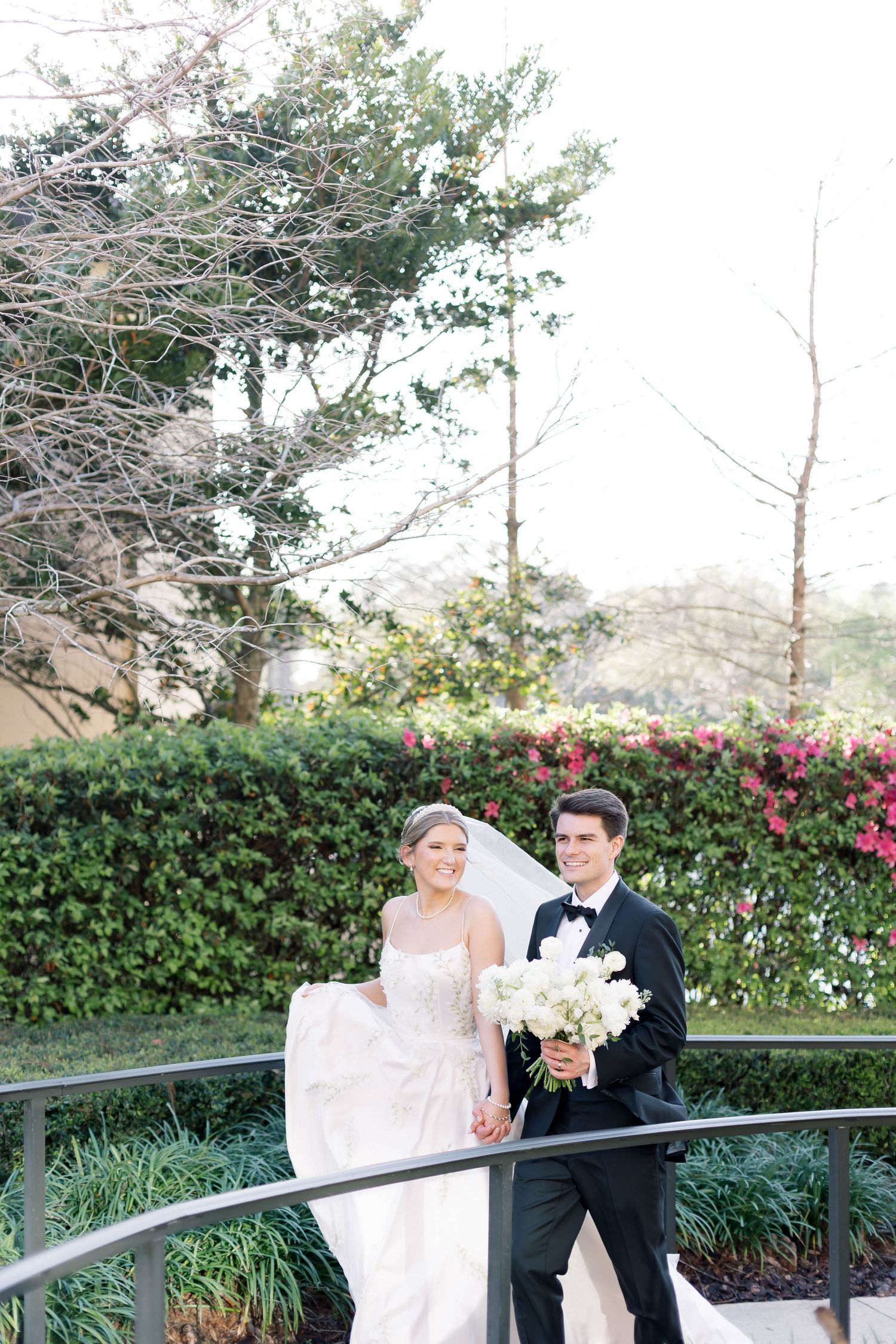 Bride and groom holding hands on a curved bridge, surrounded by lush greenery and pink flowers on their wedding day.