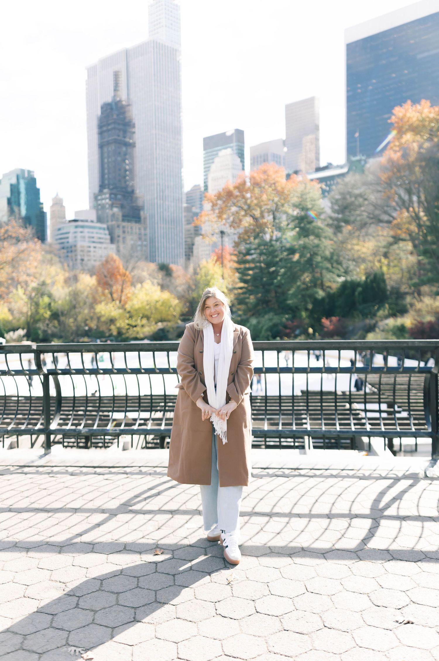 Woman in camel coat and white scarf smiling in Central Park with NYC skyline and autumn trees in background.