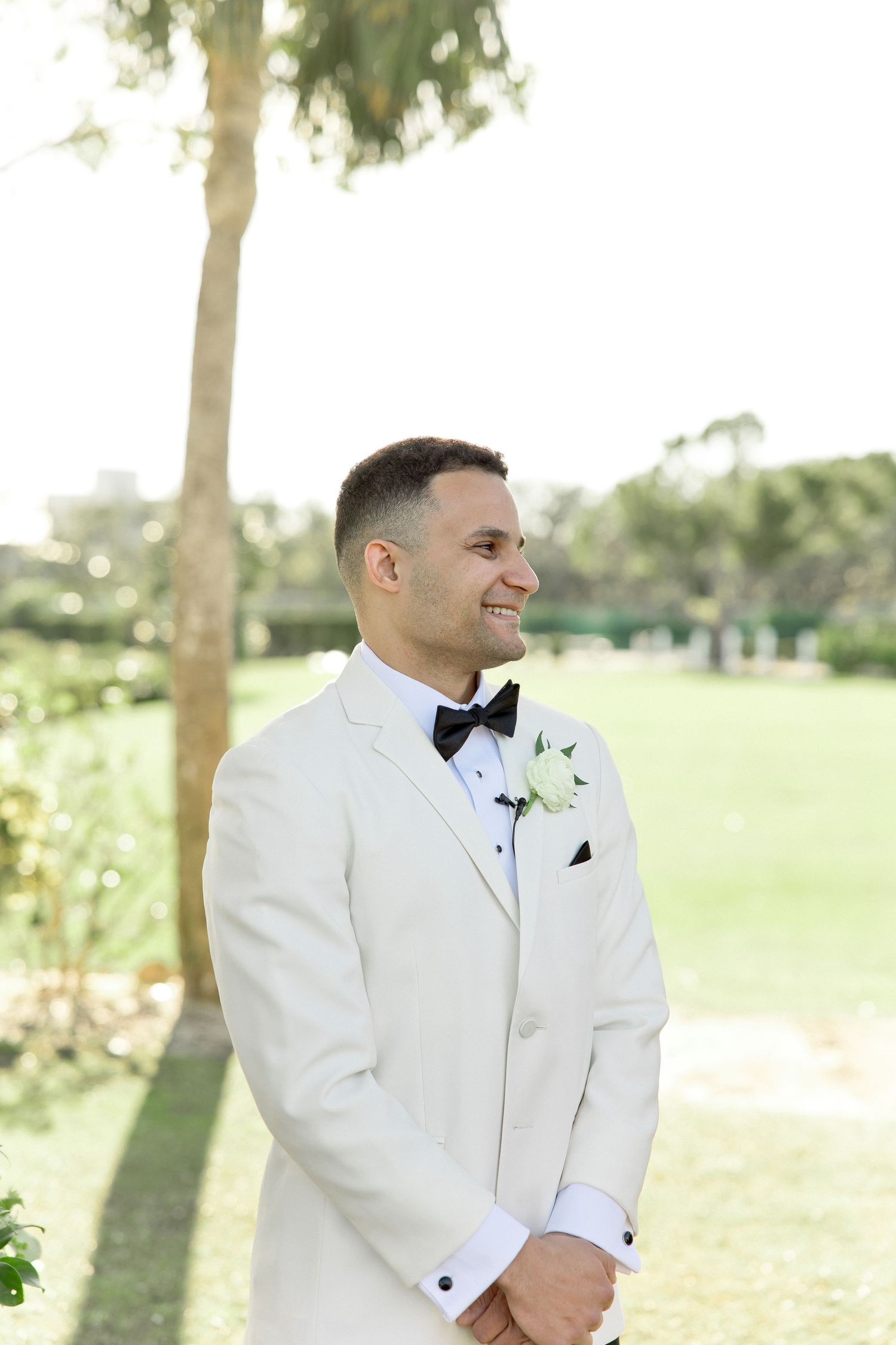 Groom in white tuxedo jacket and black bow tie smiling outdoors.
