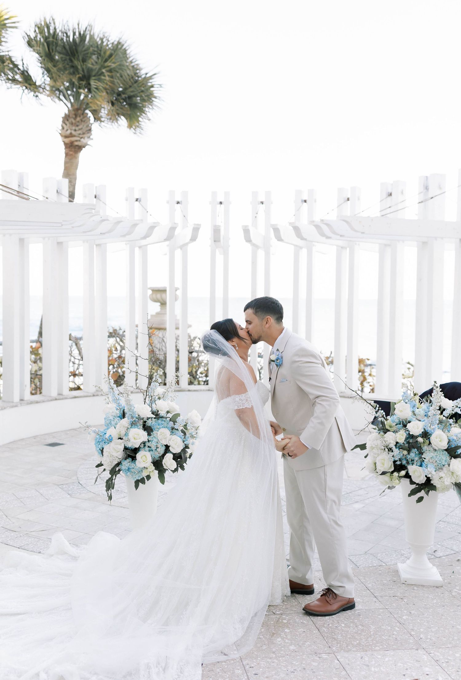 Bride and groom share first kiss at white pergola altar adorned with blue and white floral arrangements.