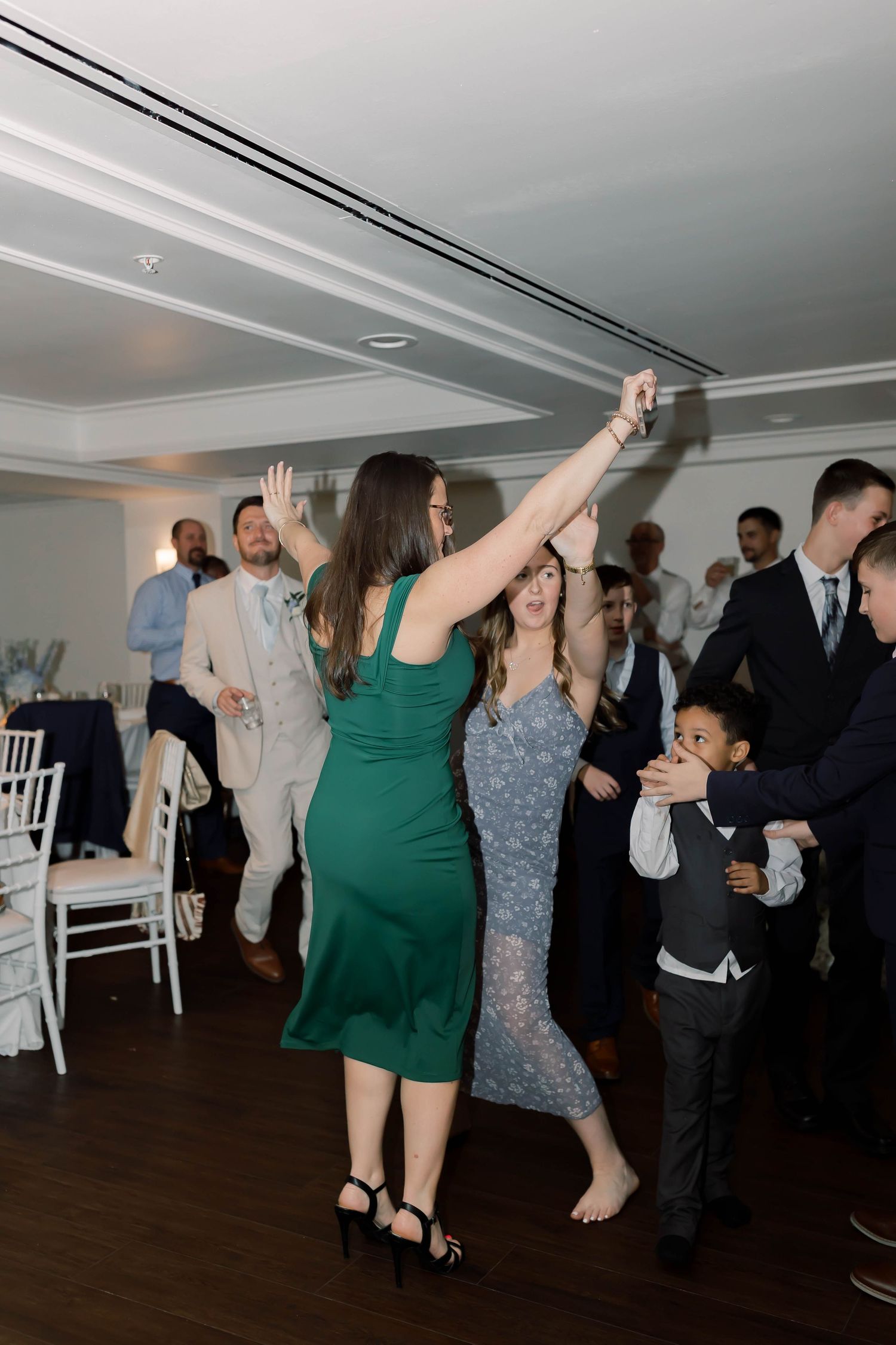 Wedding guests in formal attire dance joyfully at reception, with woman in green dress raising her hand.