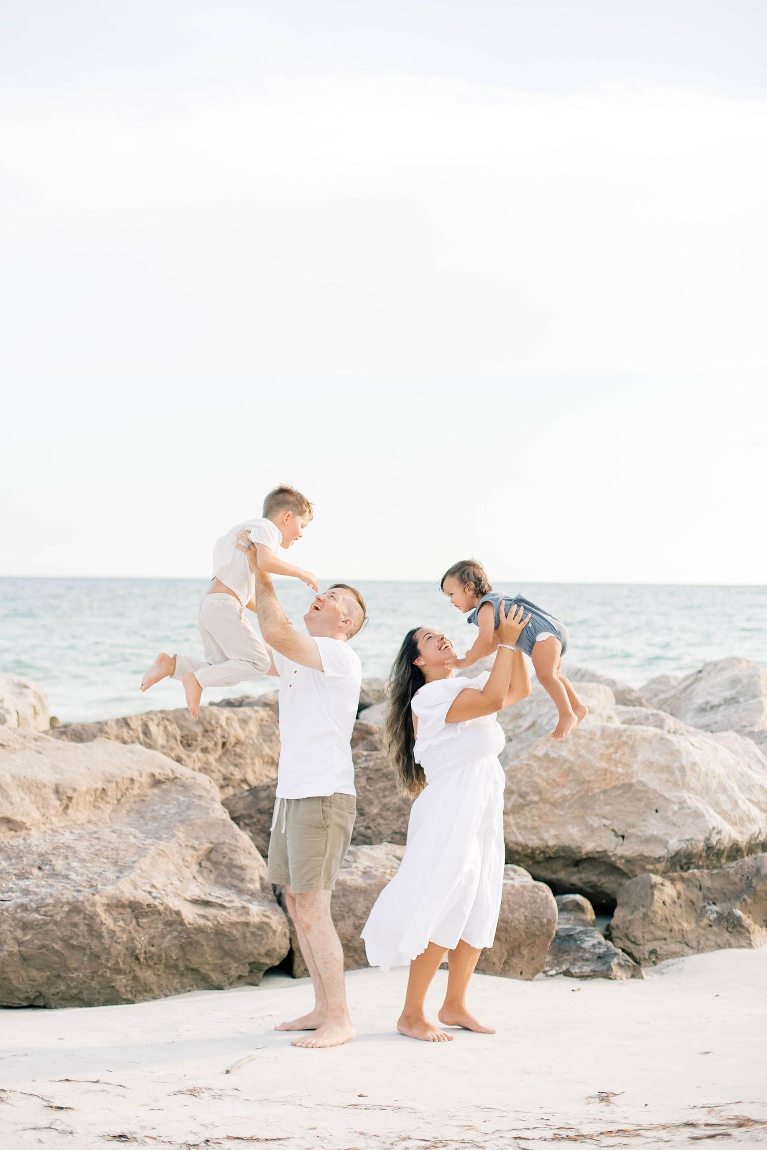 Family enjoying playful interactions on a sandy beach near large coastal rocks.