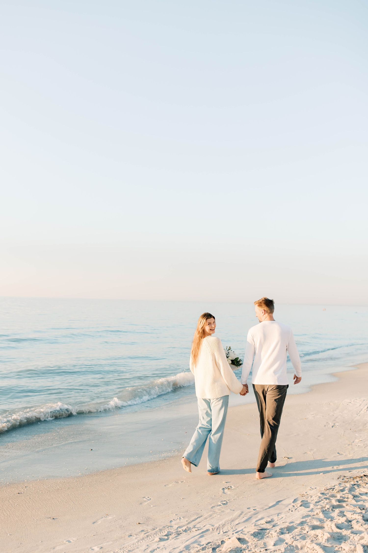A couple walks hand in hand along a pristine beach shoreline in matching white outfits.