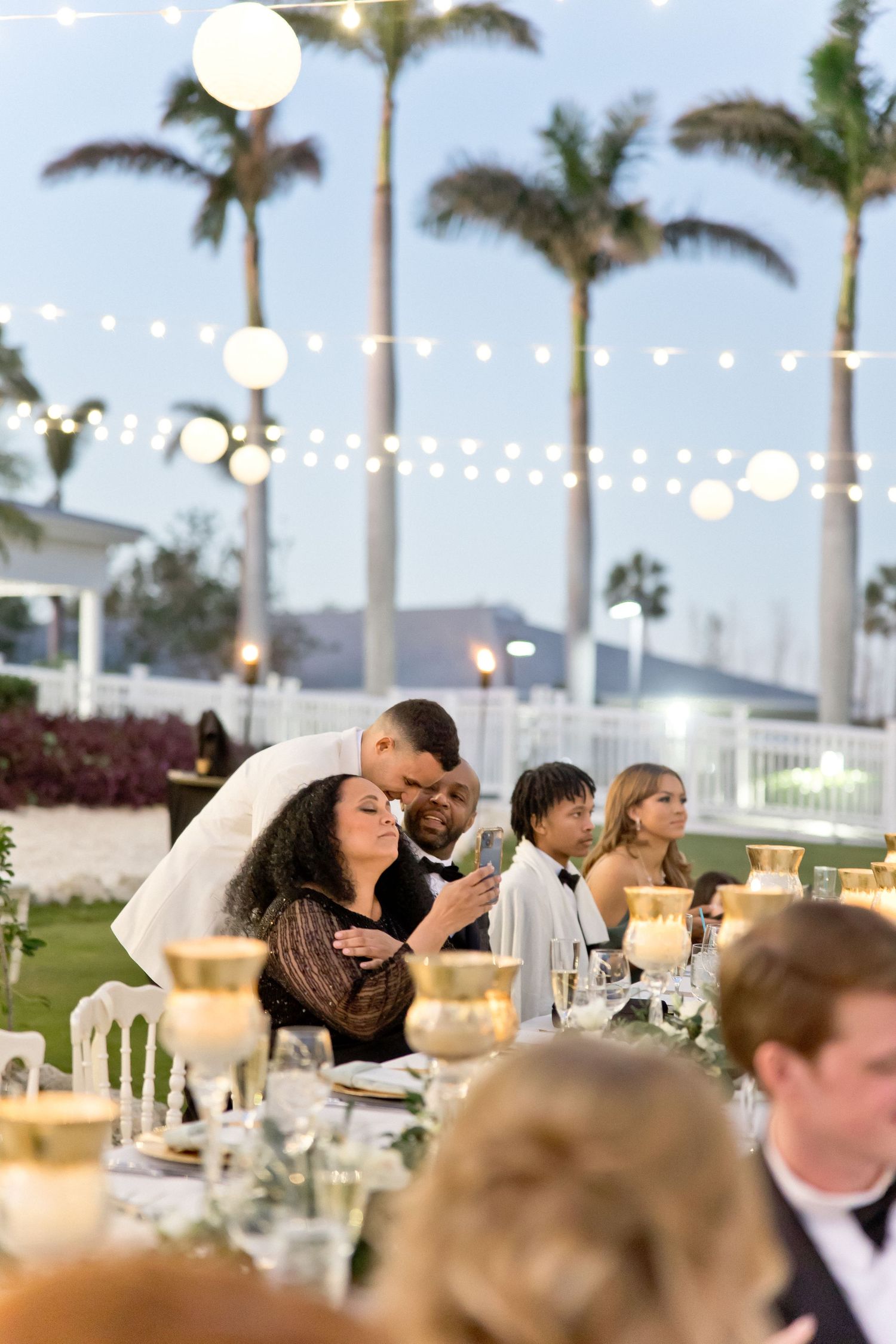 Outdoor wedding reception with guests dining under string lights and palm trees at sunset.
