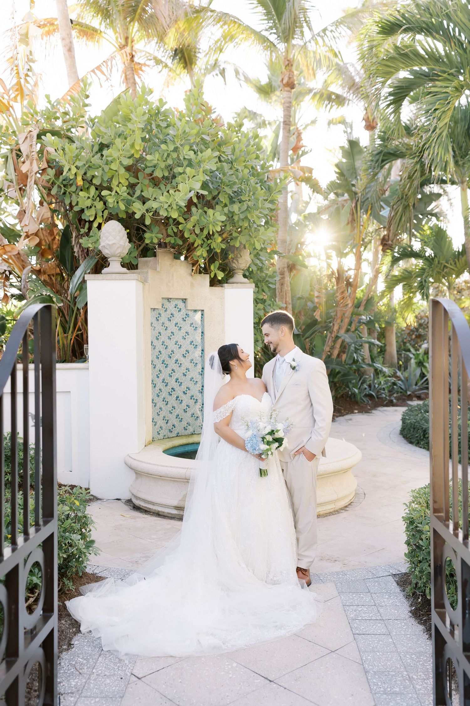 Bride and groom share a romantic moment in a lush tropical garden with a decorative fountain backdrop.