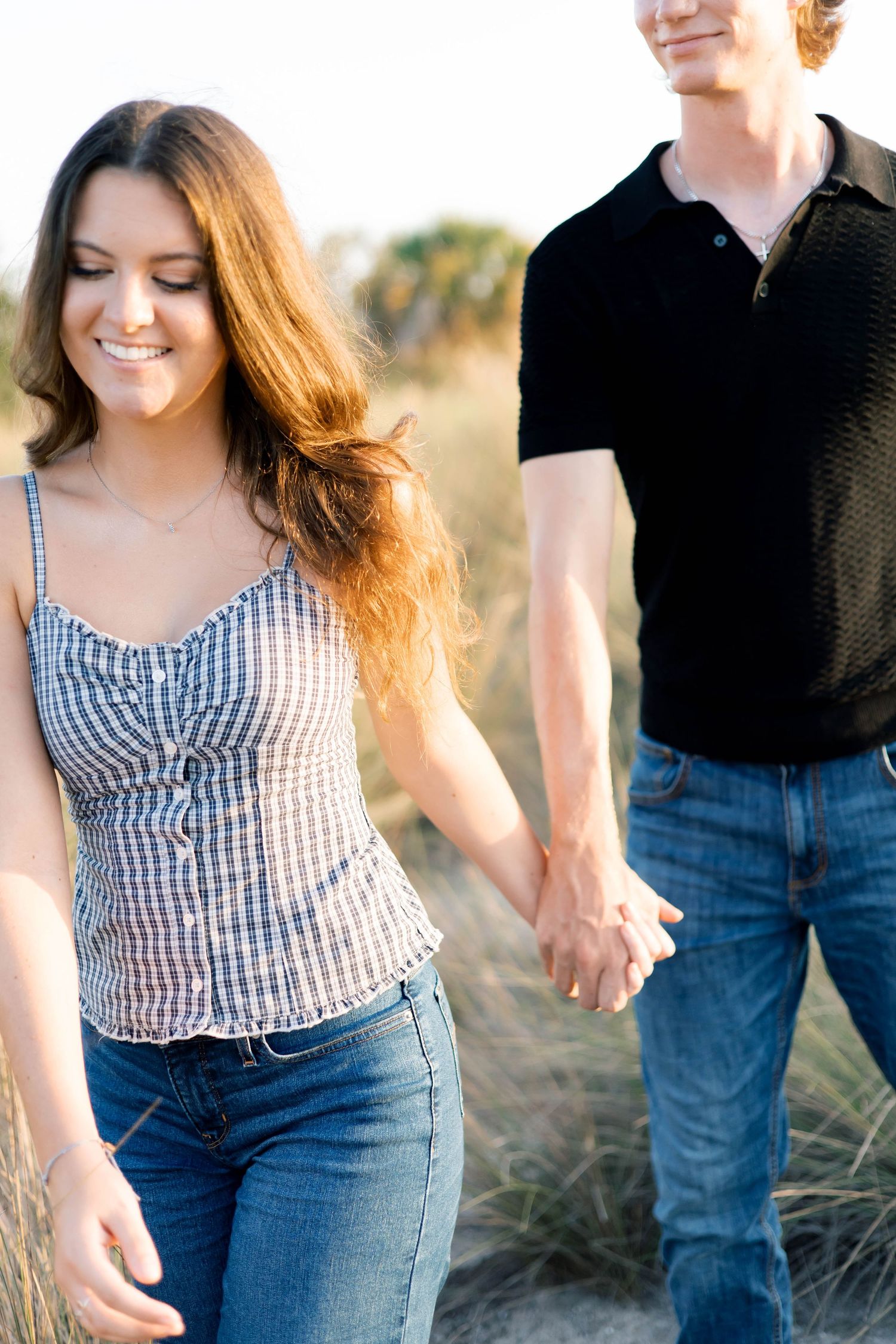 Young couple holding hands and walking outdoors on a sandy path, woman smiling in plaid top and jeans.