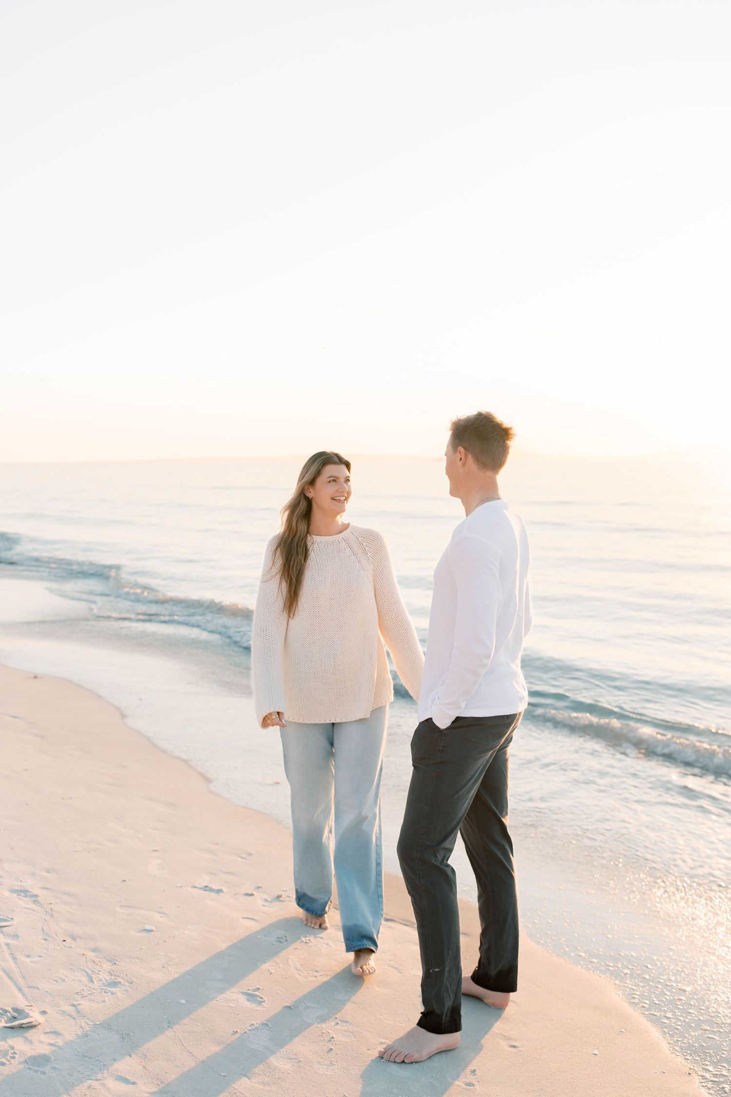 A couple strolls hand in hand along a serene beach at sunset casting long shadows.