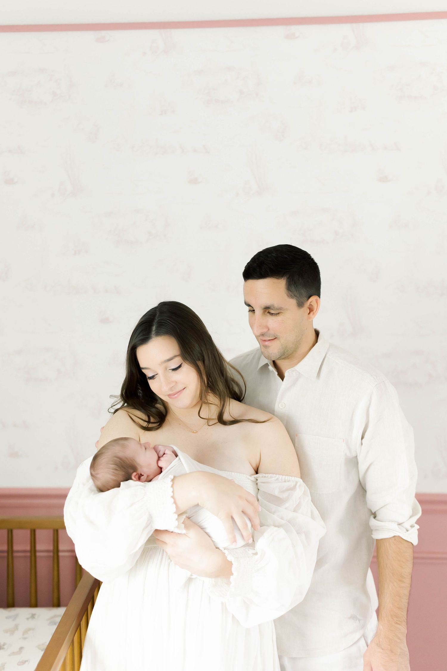Family portrait in white clothing against soft pink and white nursery walls.
