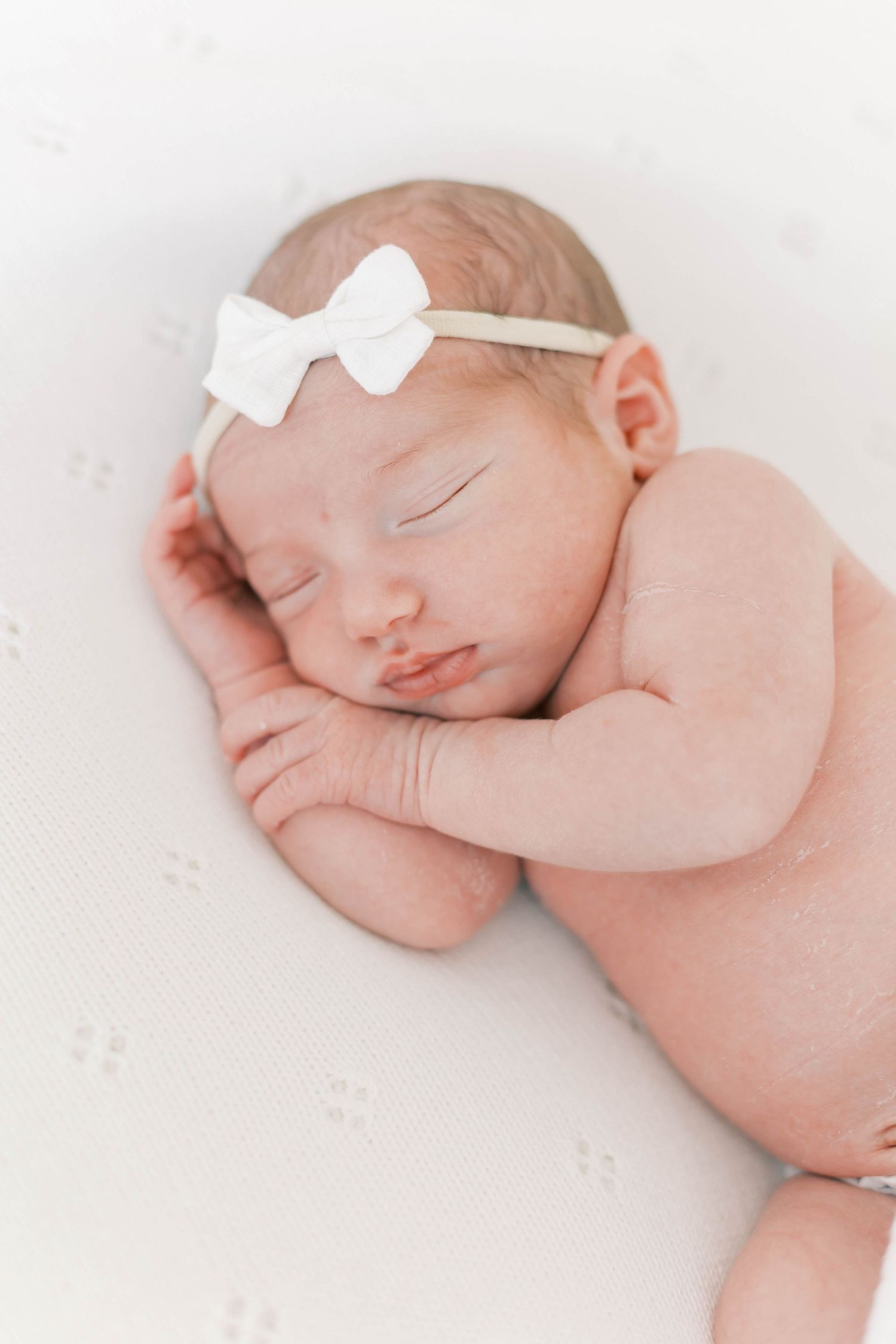 A sleeping newborn baby wearing a white bow headband rests peacefully on white fabric.
