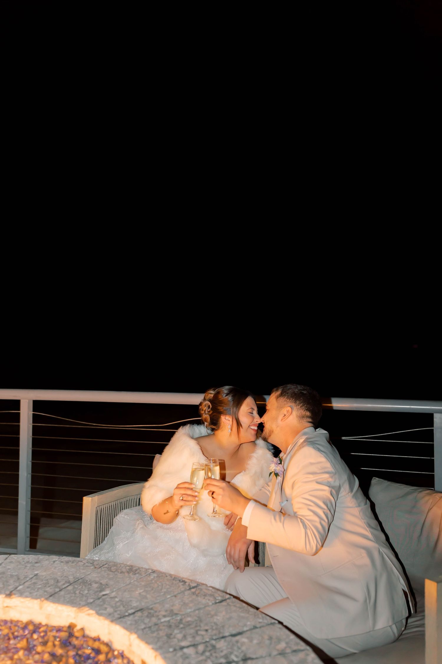 Bride and groom share a kiss while toasting champagne beside a fire pit on a dark evening.