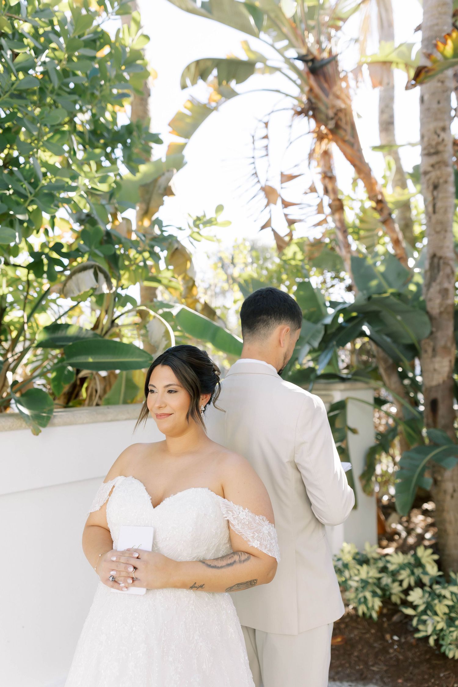 Bride in off-shoulder lace gown holds coffee cup while groom in tan suit stands behind her amid tropical greenery.