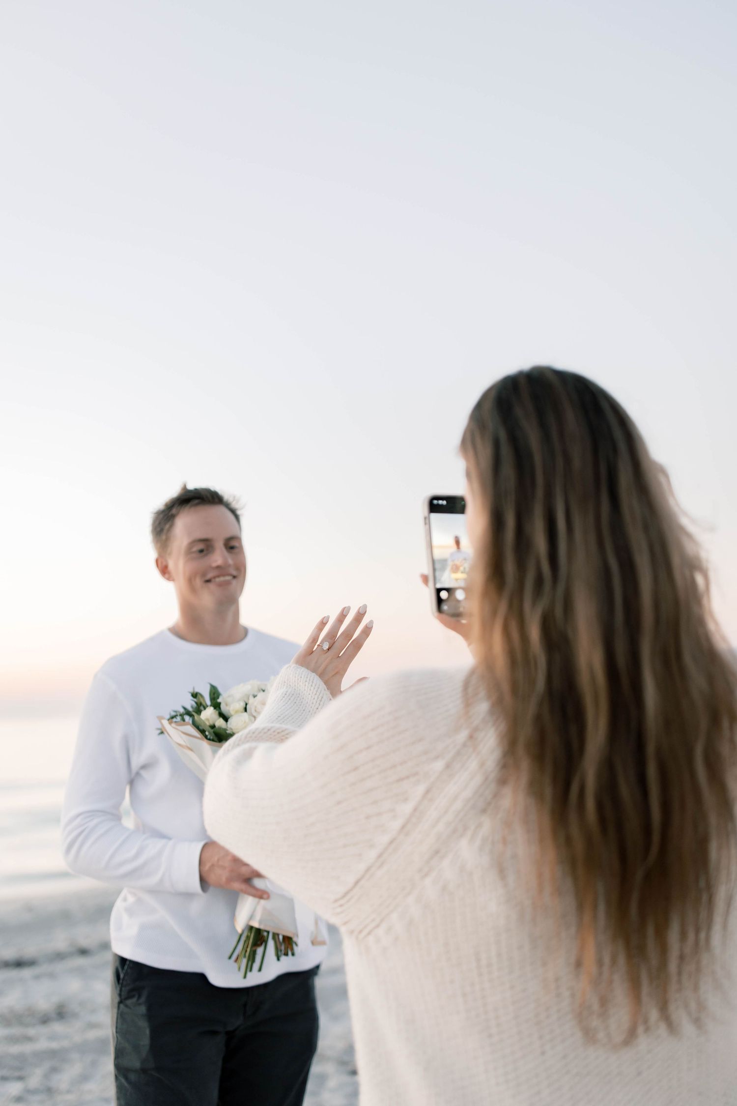 Woman photographing smiling man holding white flower bouquet on beach at sunset during romantic engagement moment.