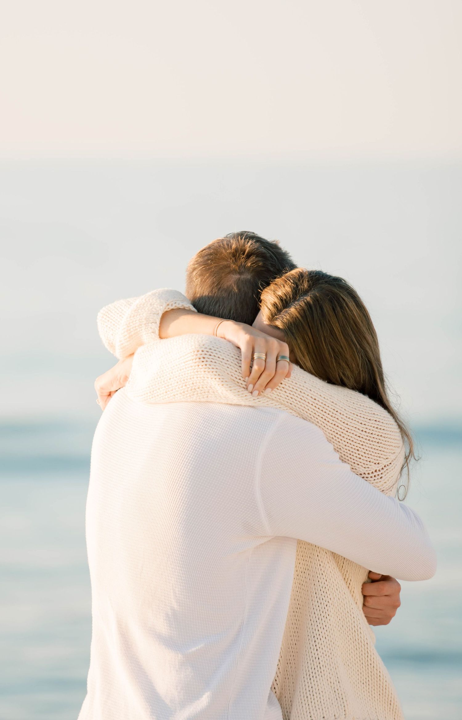 A loving couple hugs on the beach wearing matching white sweaters at sunset.