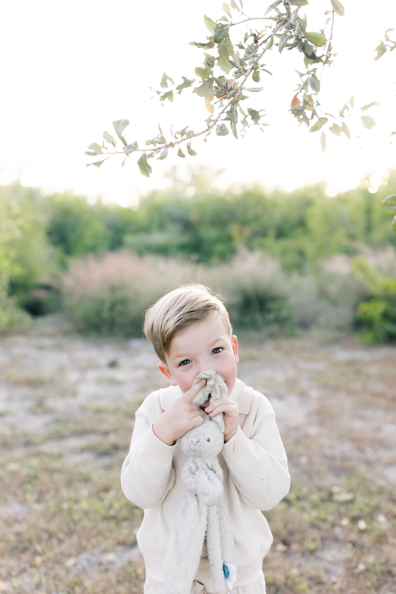 Young person in cream colored clothing holds hands to face while standing under tree branches.