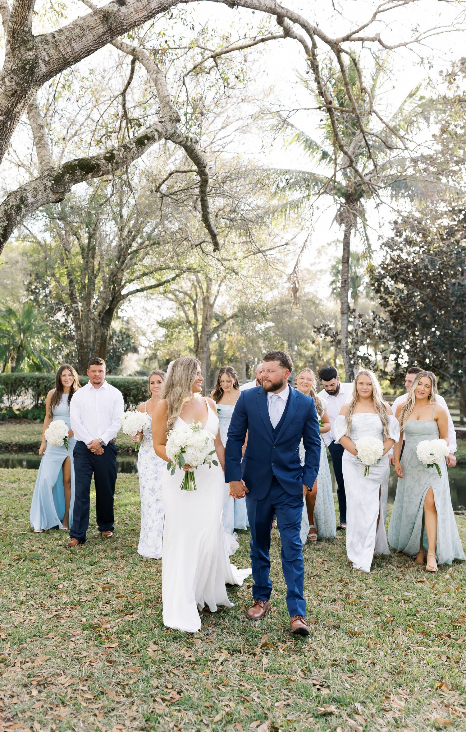 Wedding party in blue and white attire poses together on grassy lawn under trees.