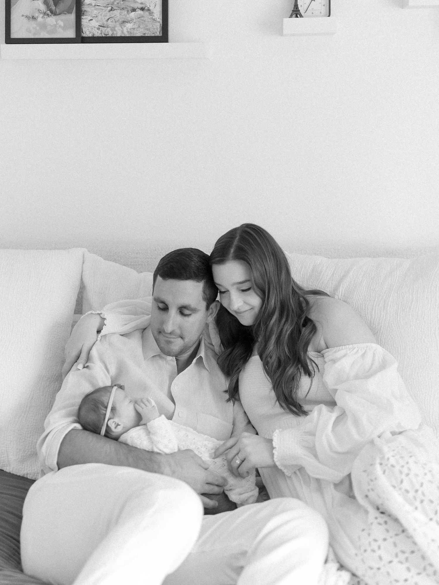 Black and white photo of parents bonding with newborn on white sofa.