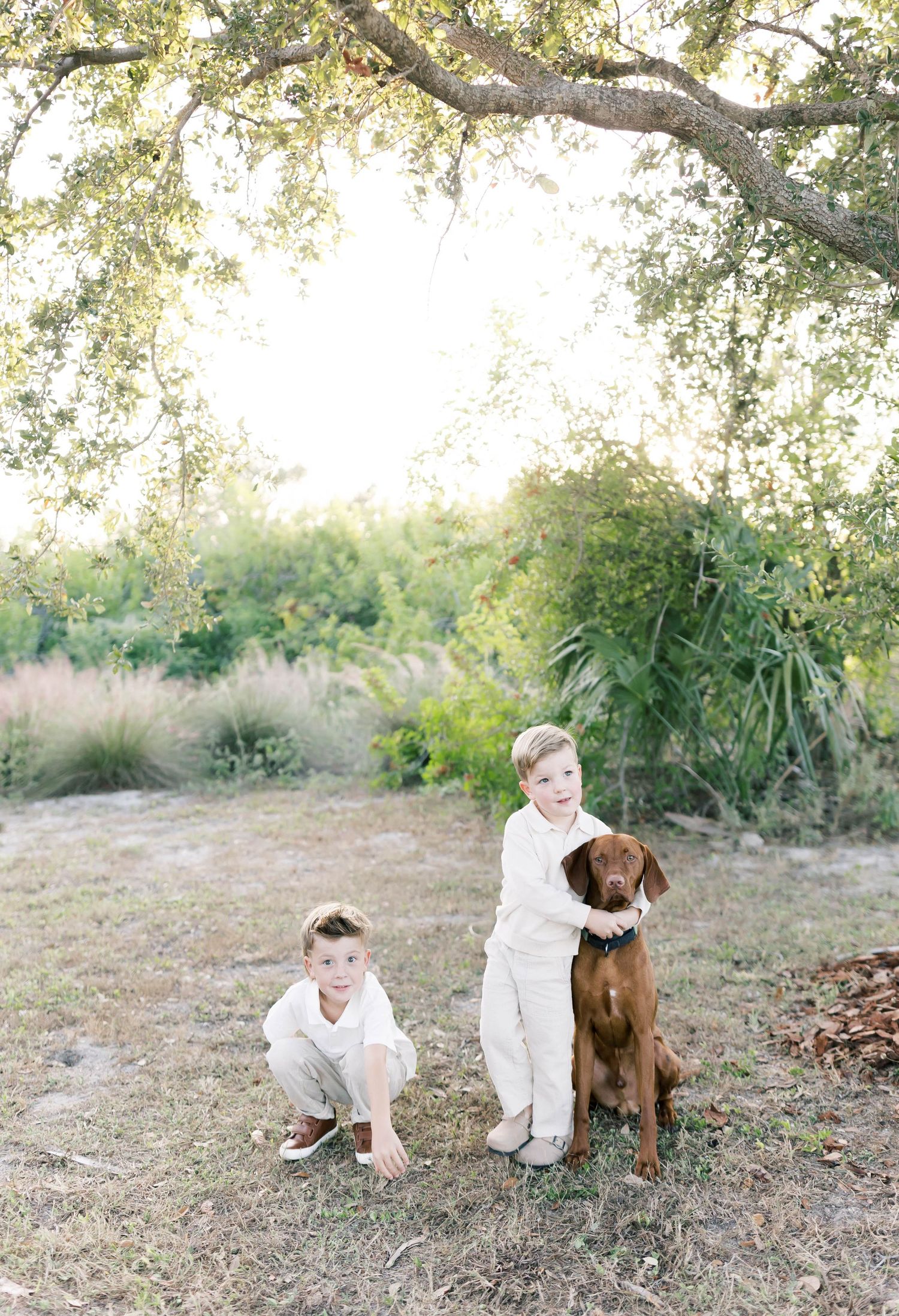Two children in white clothing pose with a brown dog in an outdoor garden setting.