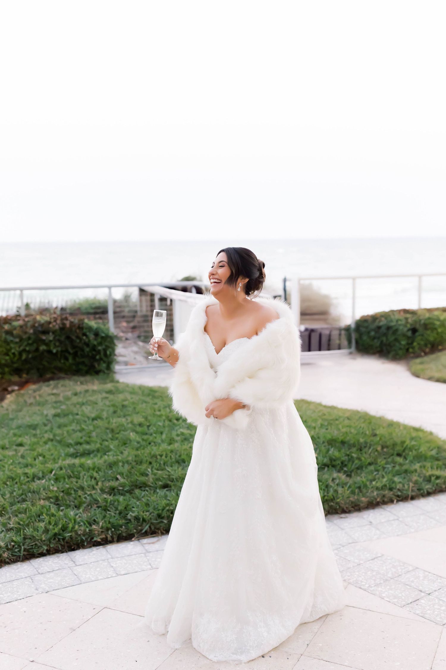 Radiant bride in white gown and fur stole laughs joyfully while holding champagne glass by the sea.
