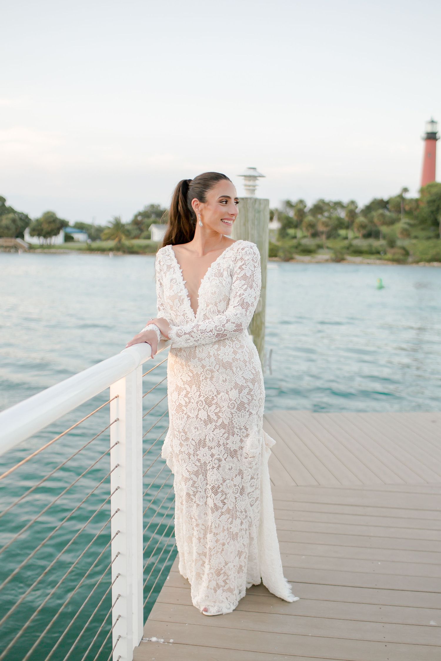 Bride in long-sleeve lace wedding gown poses on waterfront dock with lighthouse and palm trees in background.