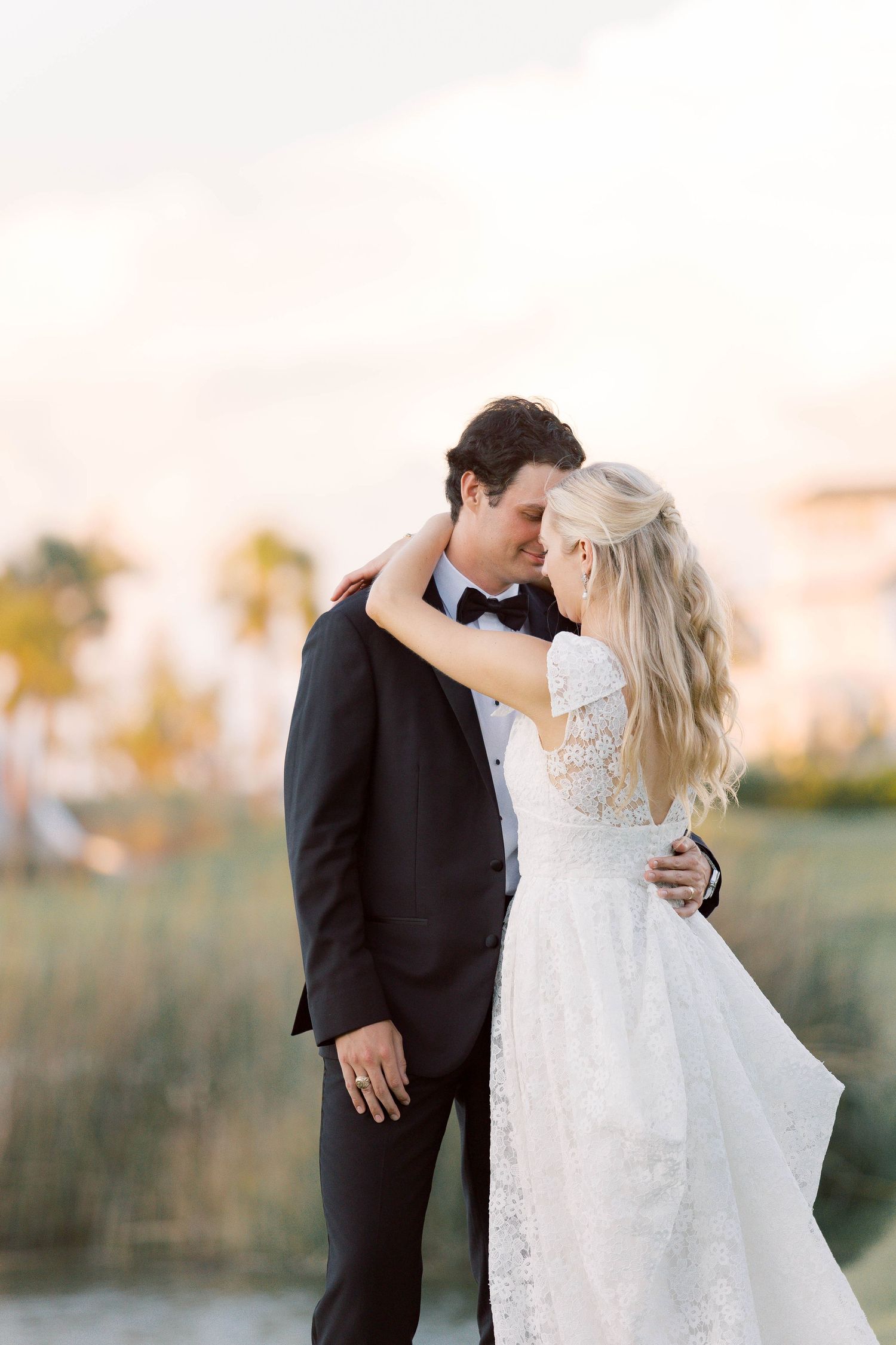 A bride and groom share a romantic embrace outdoors at golden hour, she in lace gown, he in black tuxedo.